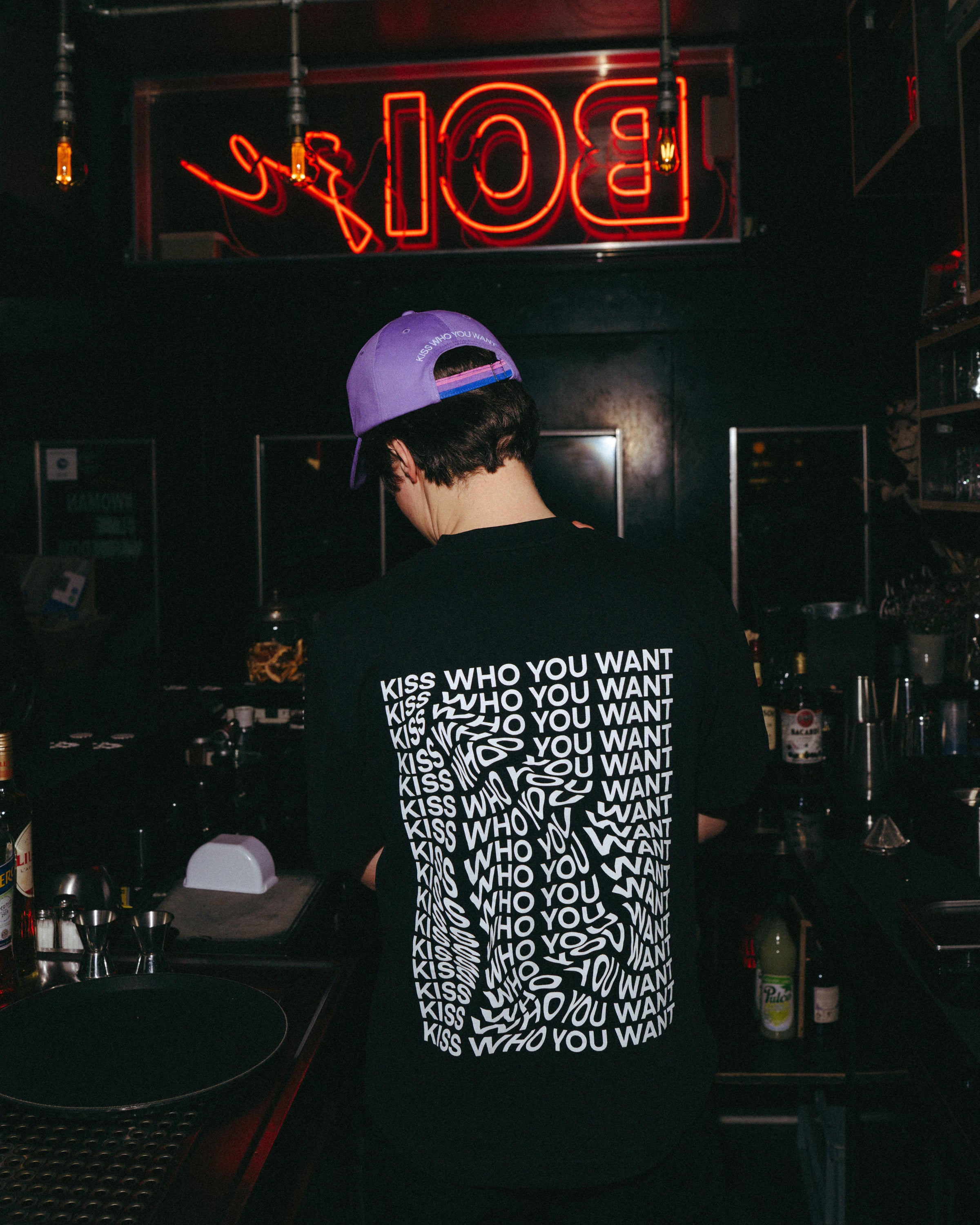 Model wearing a black oversized Biweekly T-shirt with distorted repeated ‘Kiss Who You Want’ text print, back view in a club scenario