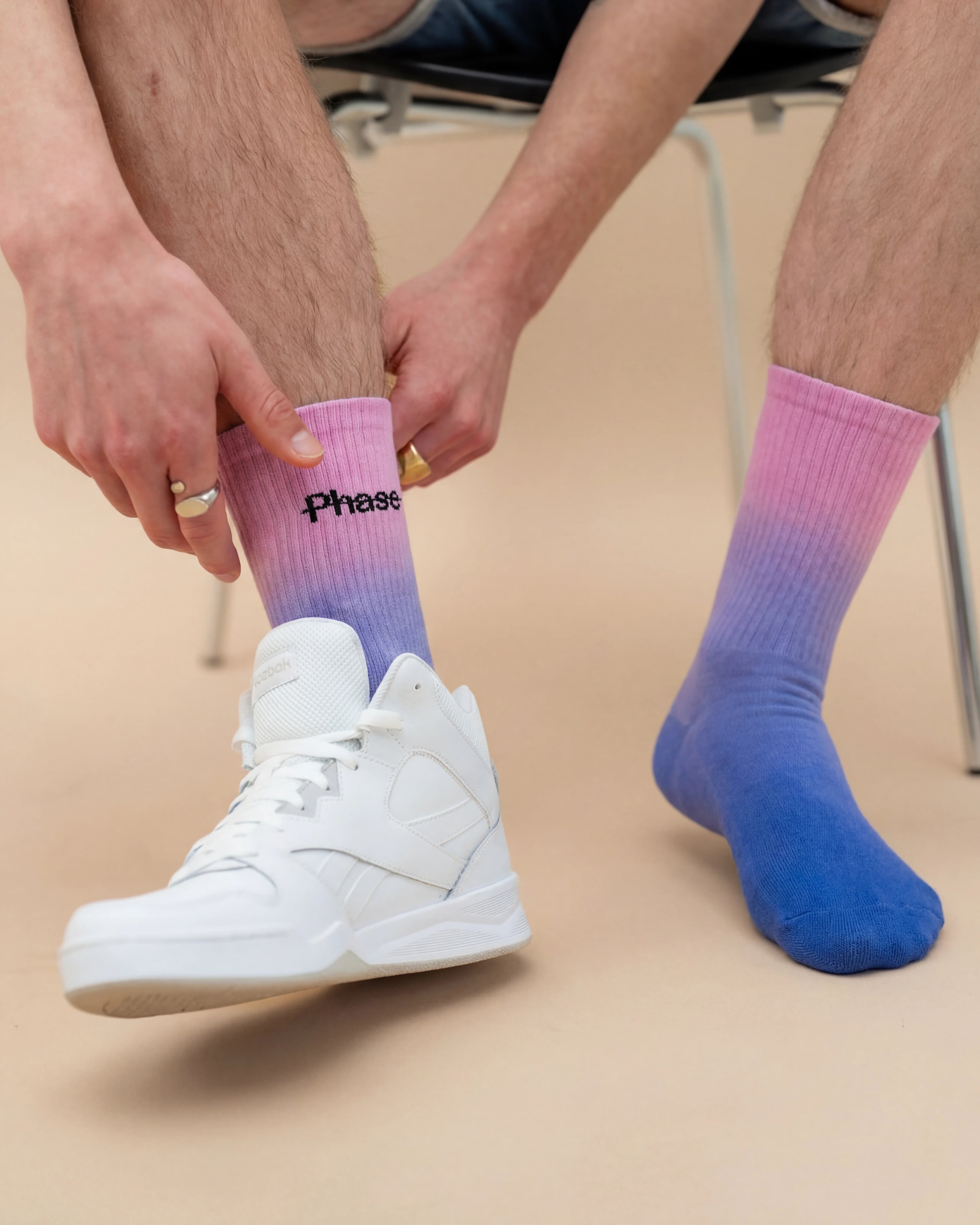 Model wearing Biweekly socks with watercolor fade from pink to blue with a crossed out "Phase" on the inside, paired with white sneakers, against a beige background
