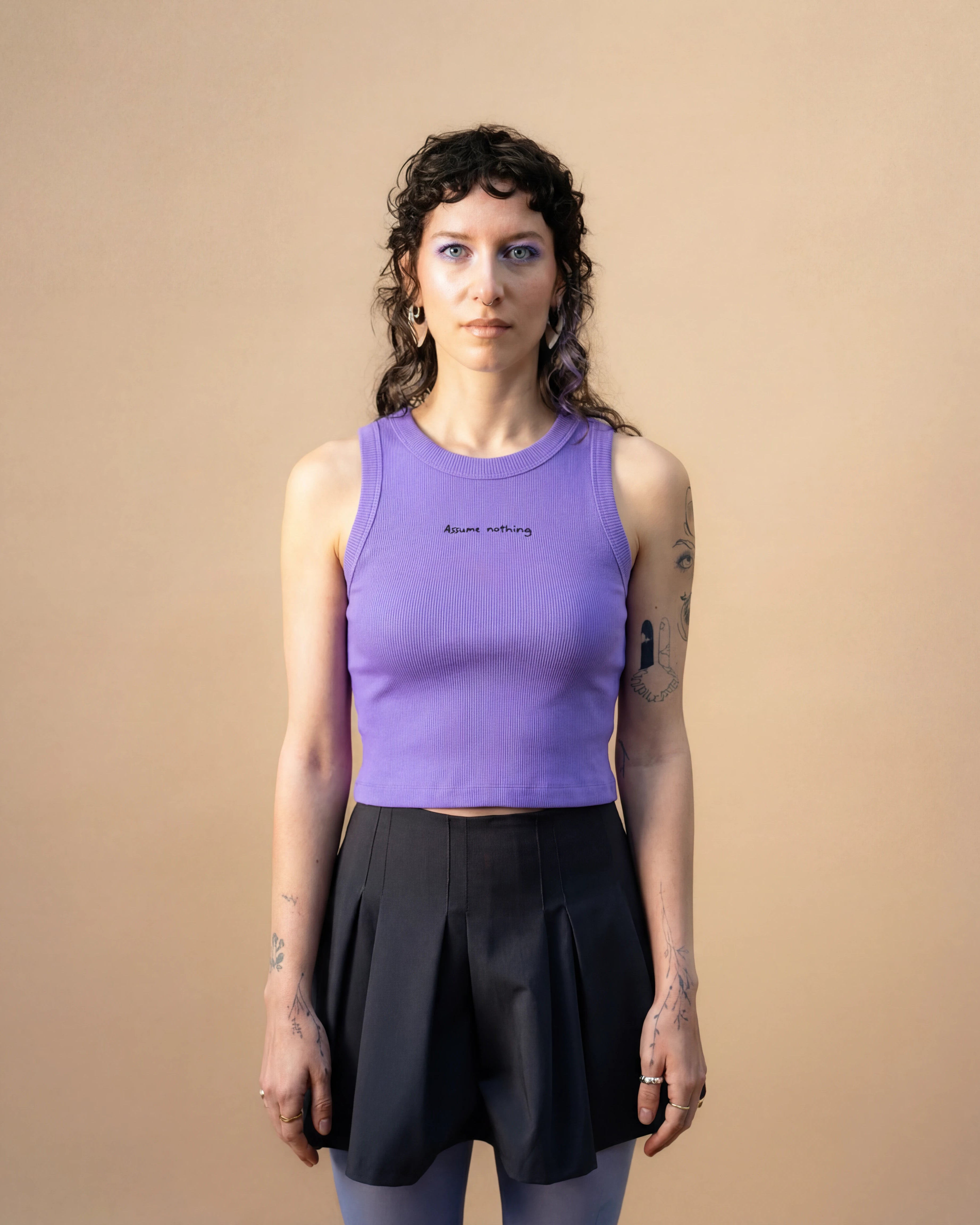 Model wearing a lilac Biweekly tank top with black ‘Assume nothing’ text, front view against a beige background
