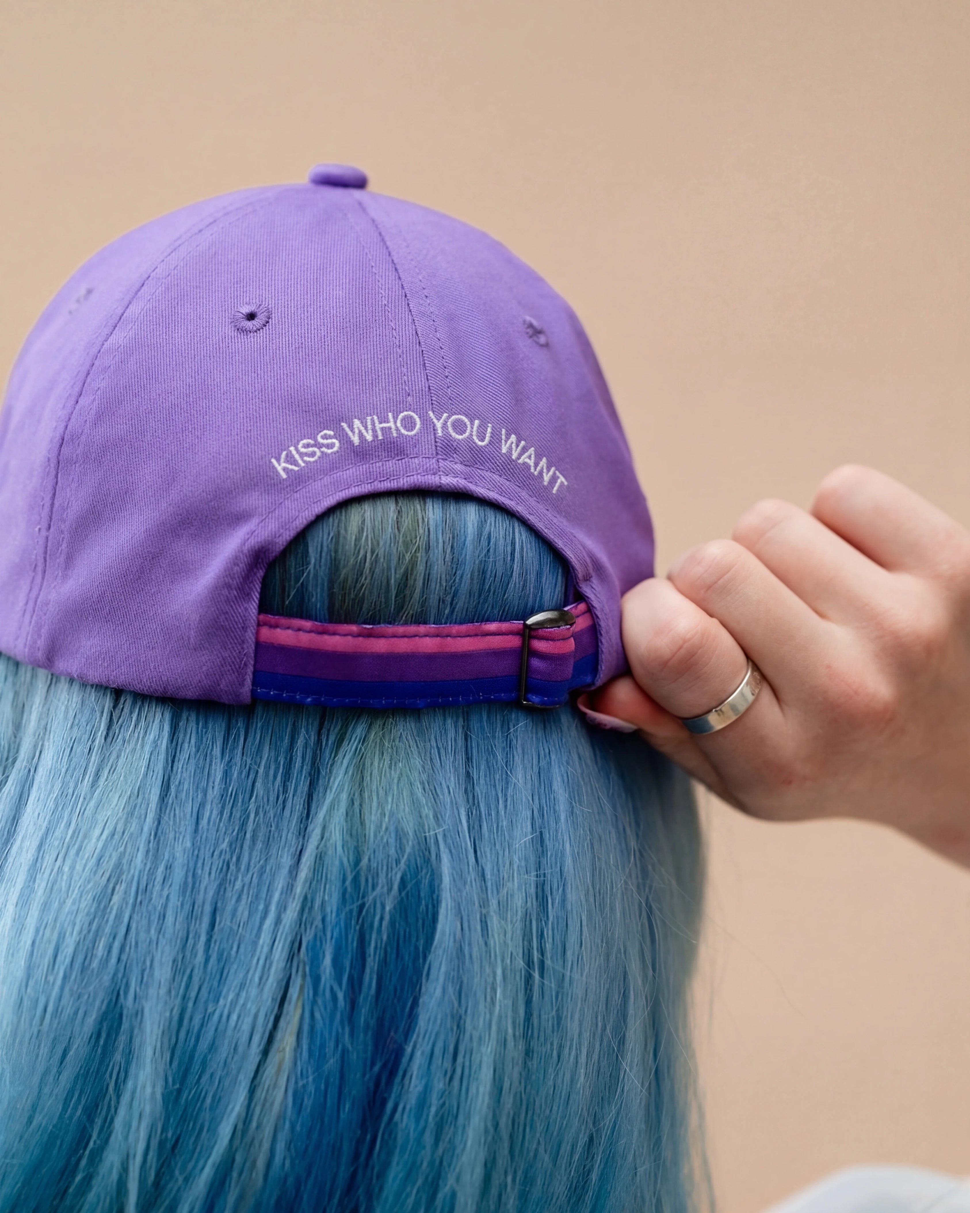 Detailshot of model wearing a lilac Biweekly cap with a strap in the colors of the bi pride flag and the text ‘Kiss Who You Want’ on the back, back view, against a beige background