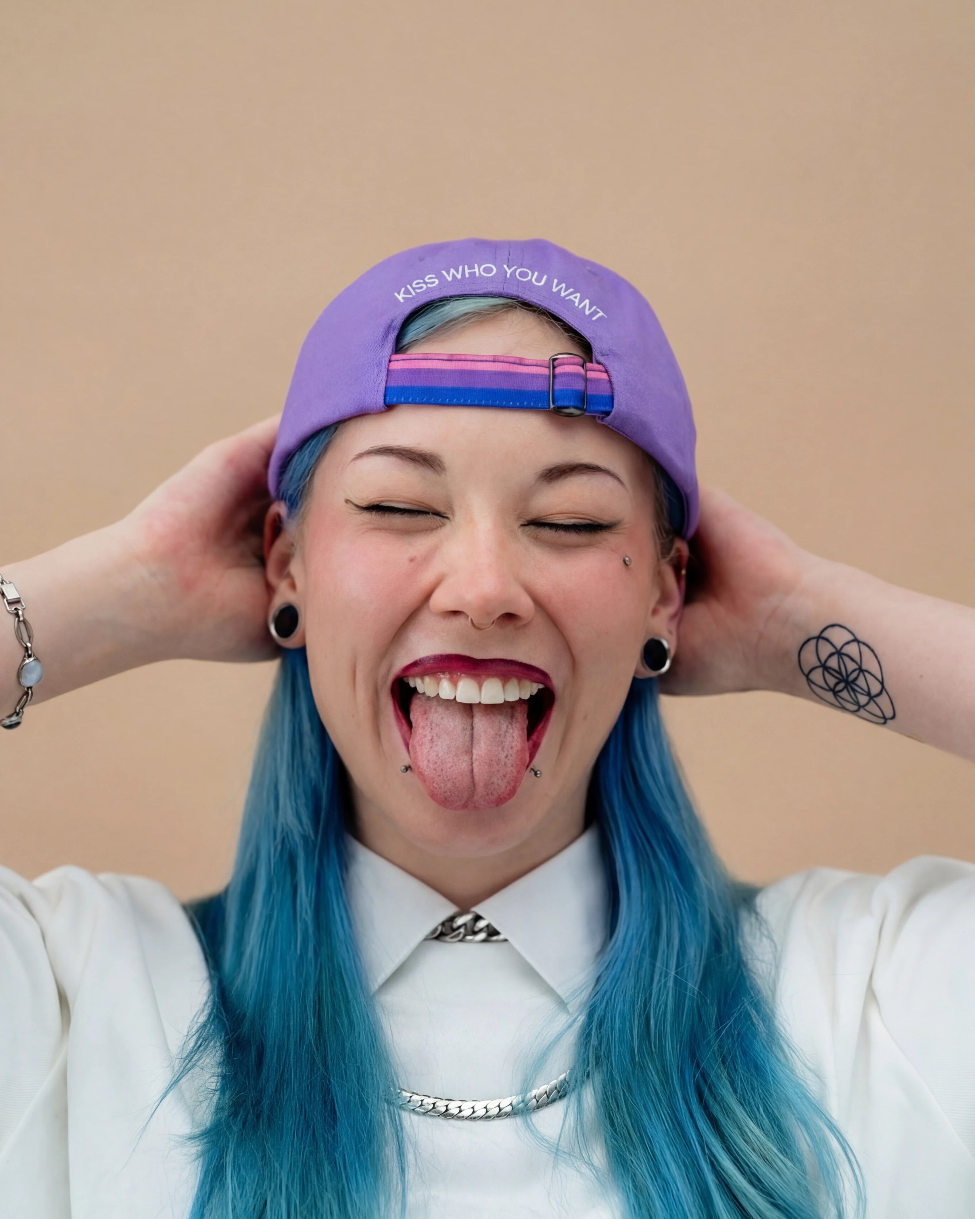 Model wearing a lilac Biweekly cap with a strap in the colors of the bi pride flag and the text ‘Kiss Who You Want’ on the back, sticking out their tongue against a beige background