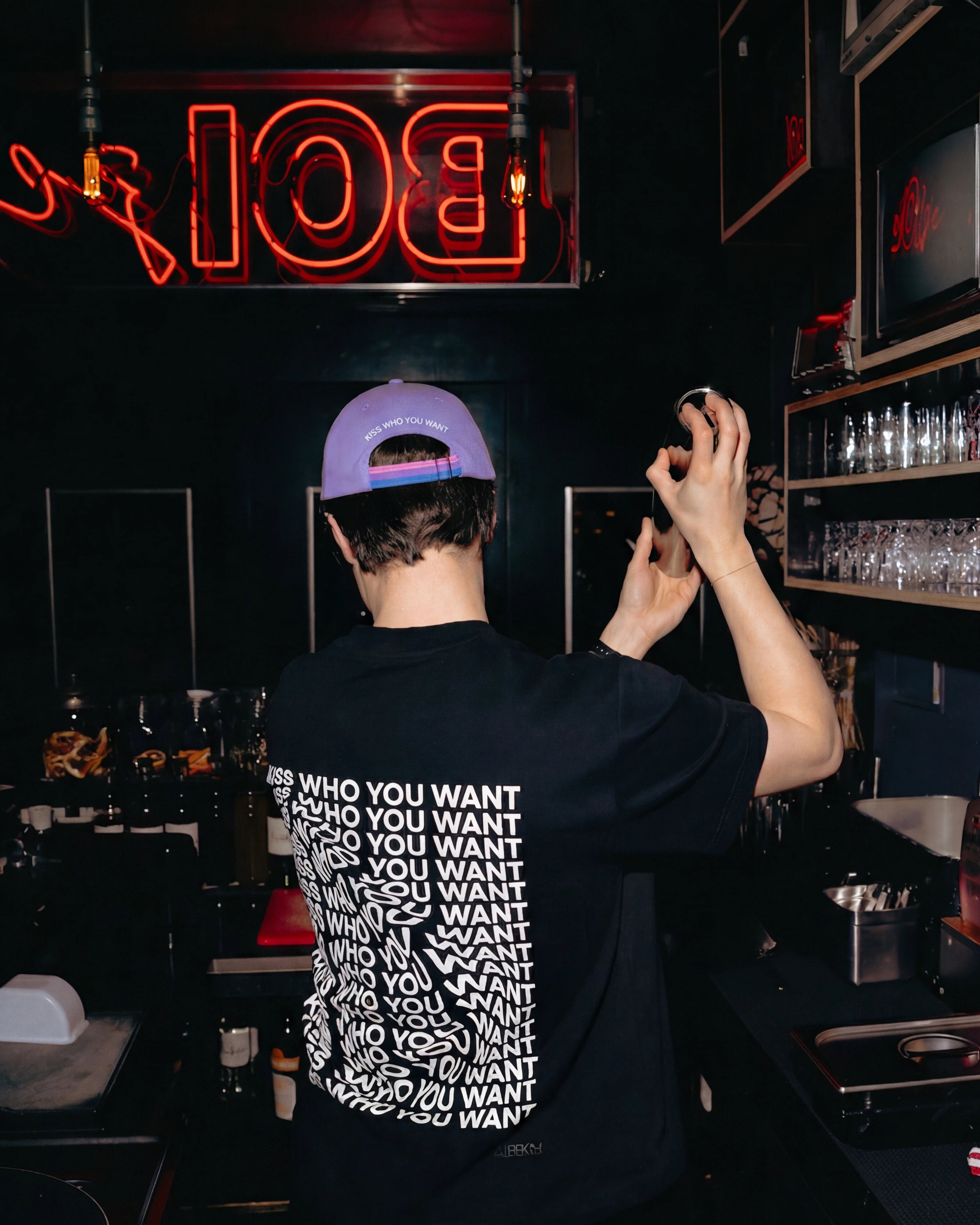 Model wearing a lilac Biweekly cap with bi pride flag strap and ‘Kiss Who You Want’ text on the back, back view, in a club scenario with a cocktail shaker in his hand