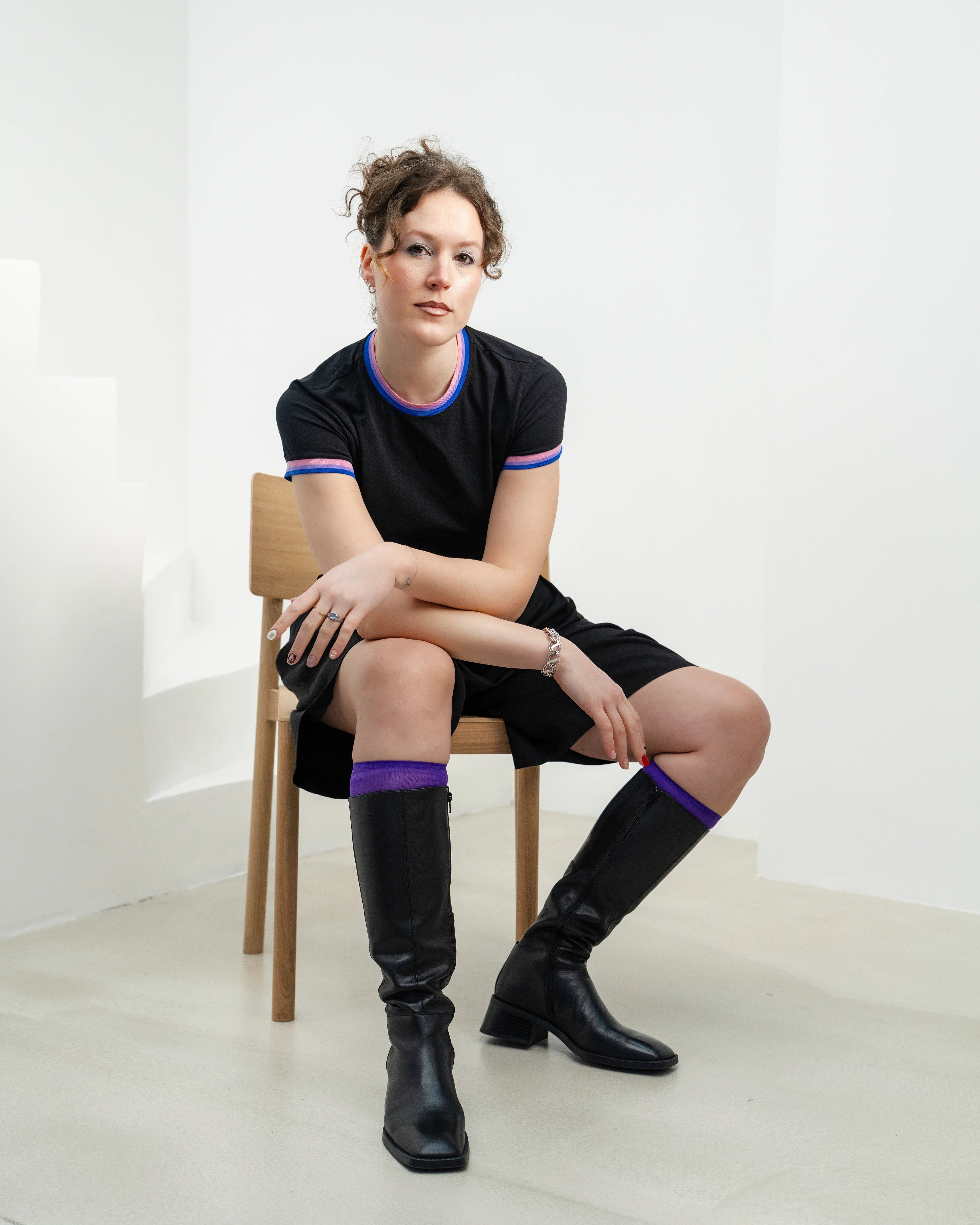 Model wearing a black Biweekly Babytee with bi pride flag trim on the collar and sleeves and black logo in the front, sitting on a chair front view against a white background