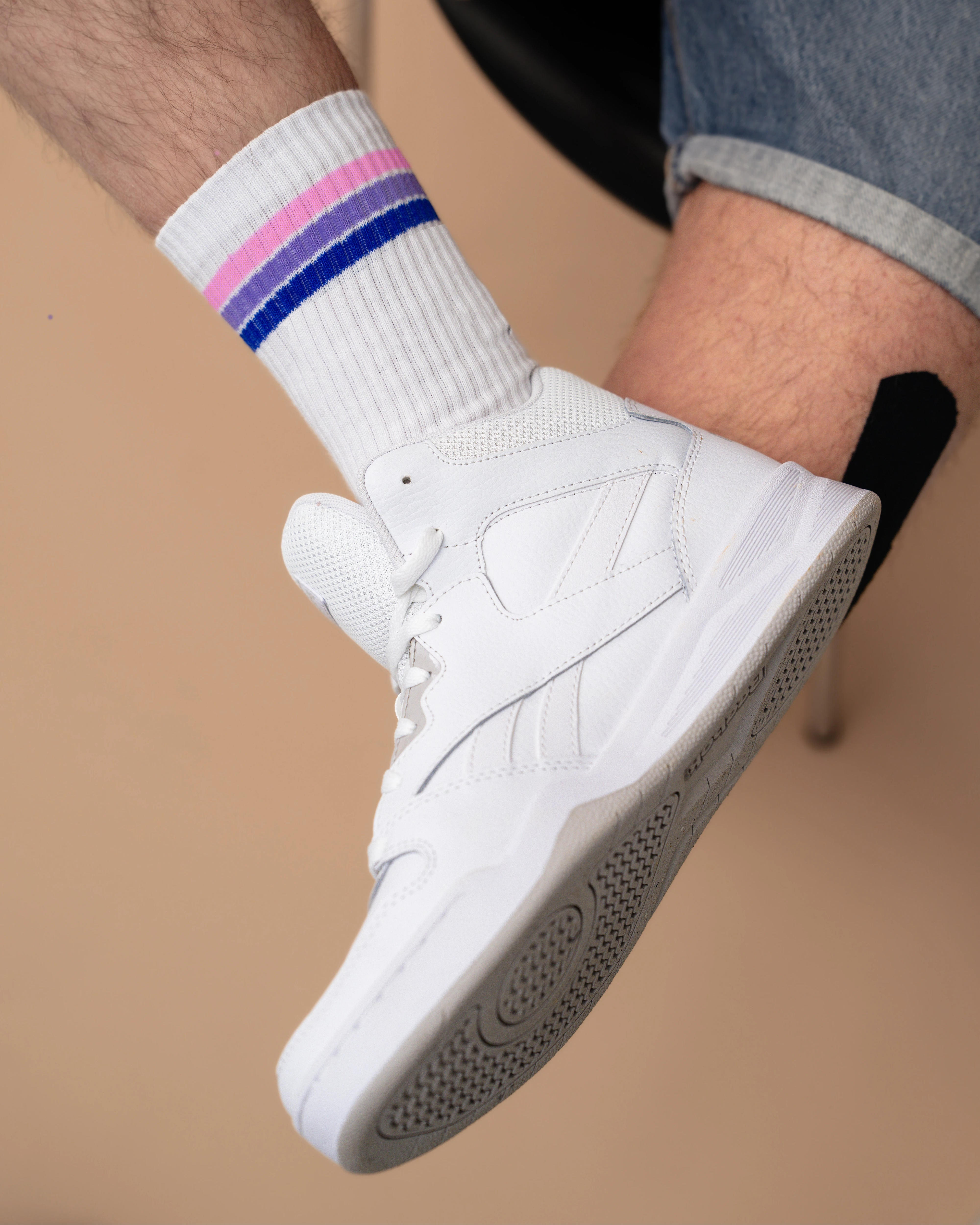 Detailshot of white Biweekly socks with bi pride flag stripes, paired with white sneakers, against a beige background
