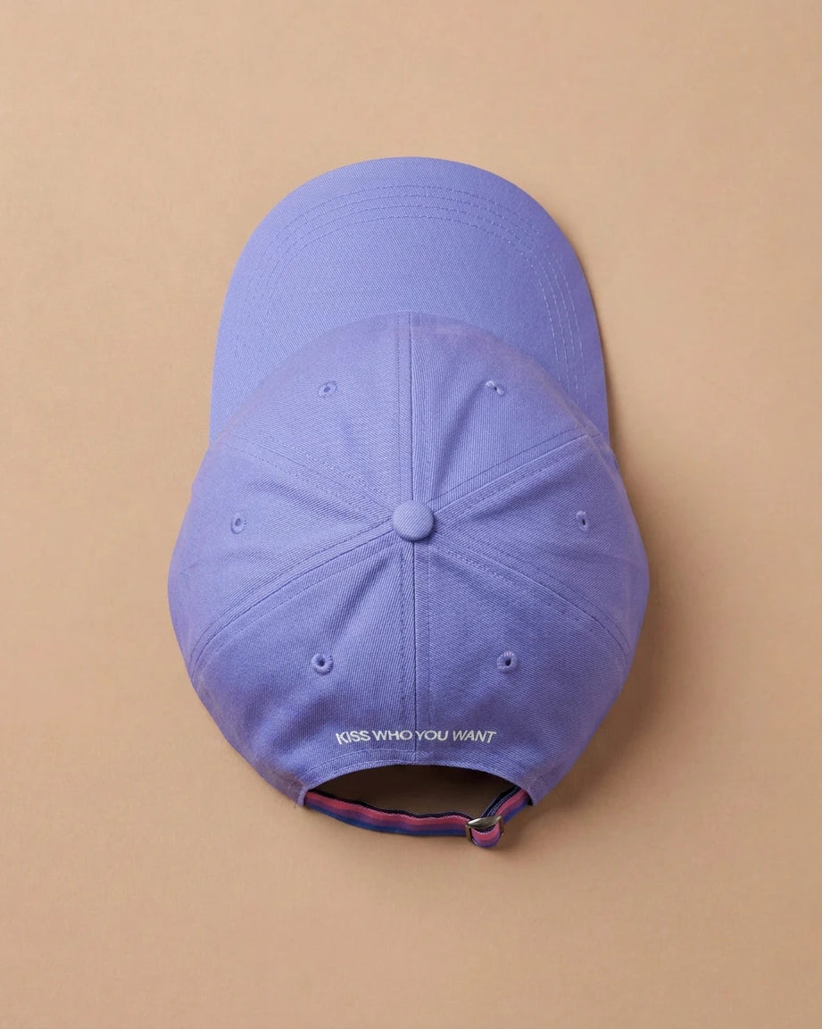 Lilac Biweekly cap with a strap in the colors of the bi pride flag and the text ‘Kiss Who You Want’ on the back, laying flat on beige background