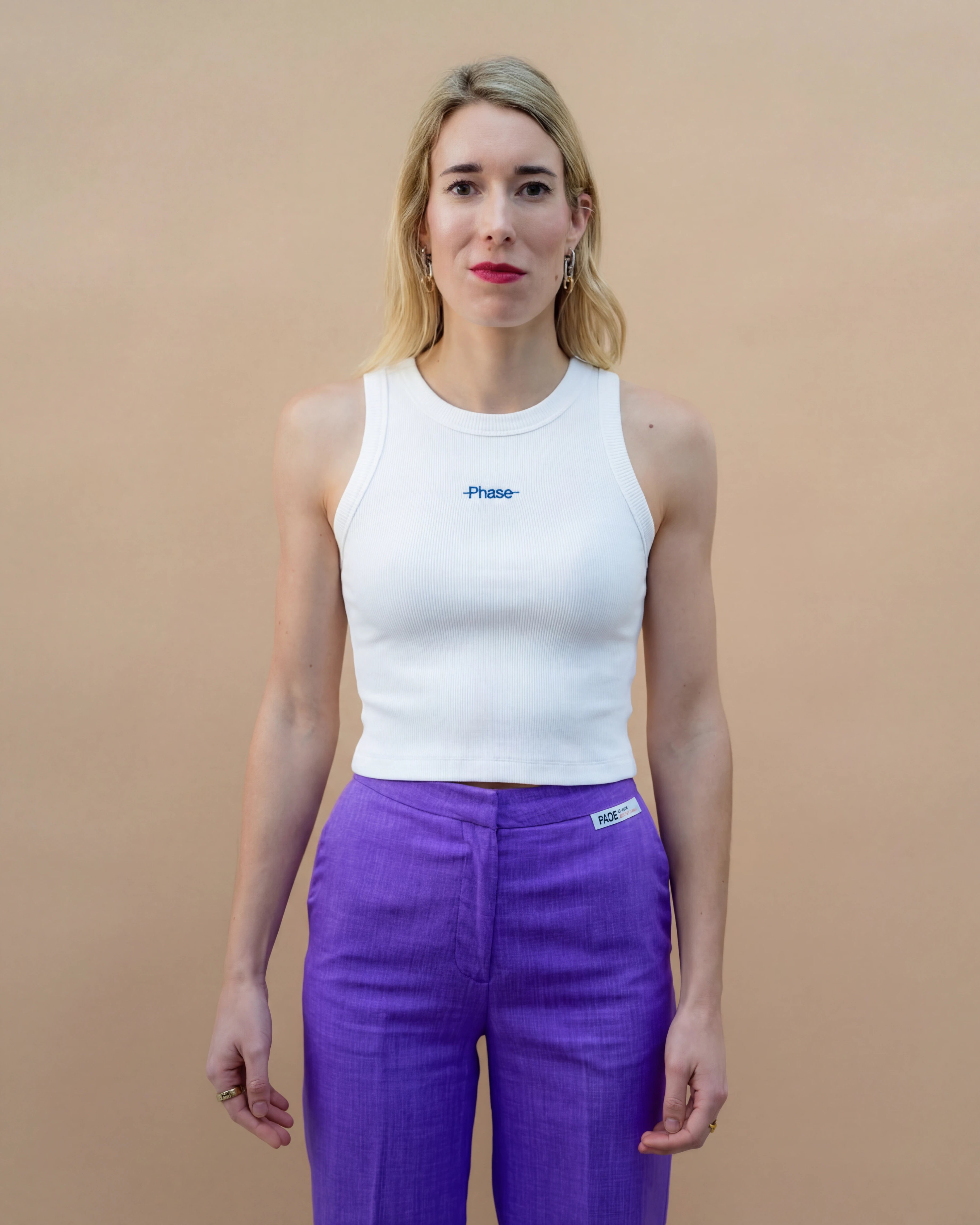 Model wearing a white Biweekly tank top with blue ‘Phase’ text that is crossed out, front view against a beige background