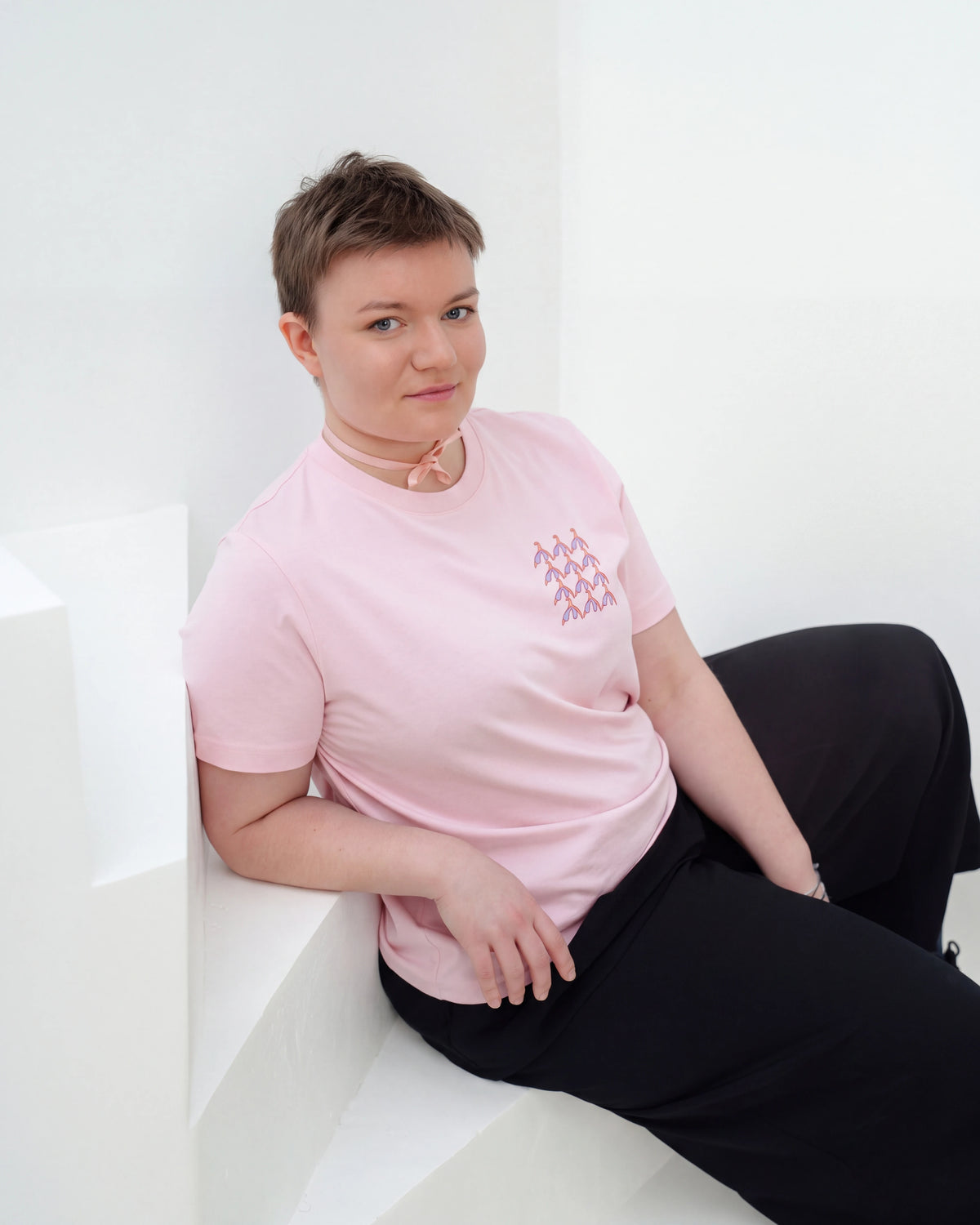 Model wearing light pink Biweekly T-shirt with clit graphic print on right chest area, front view, sitting on stairs against white background
