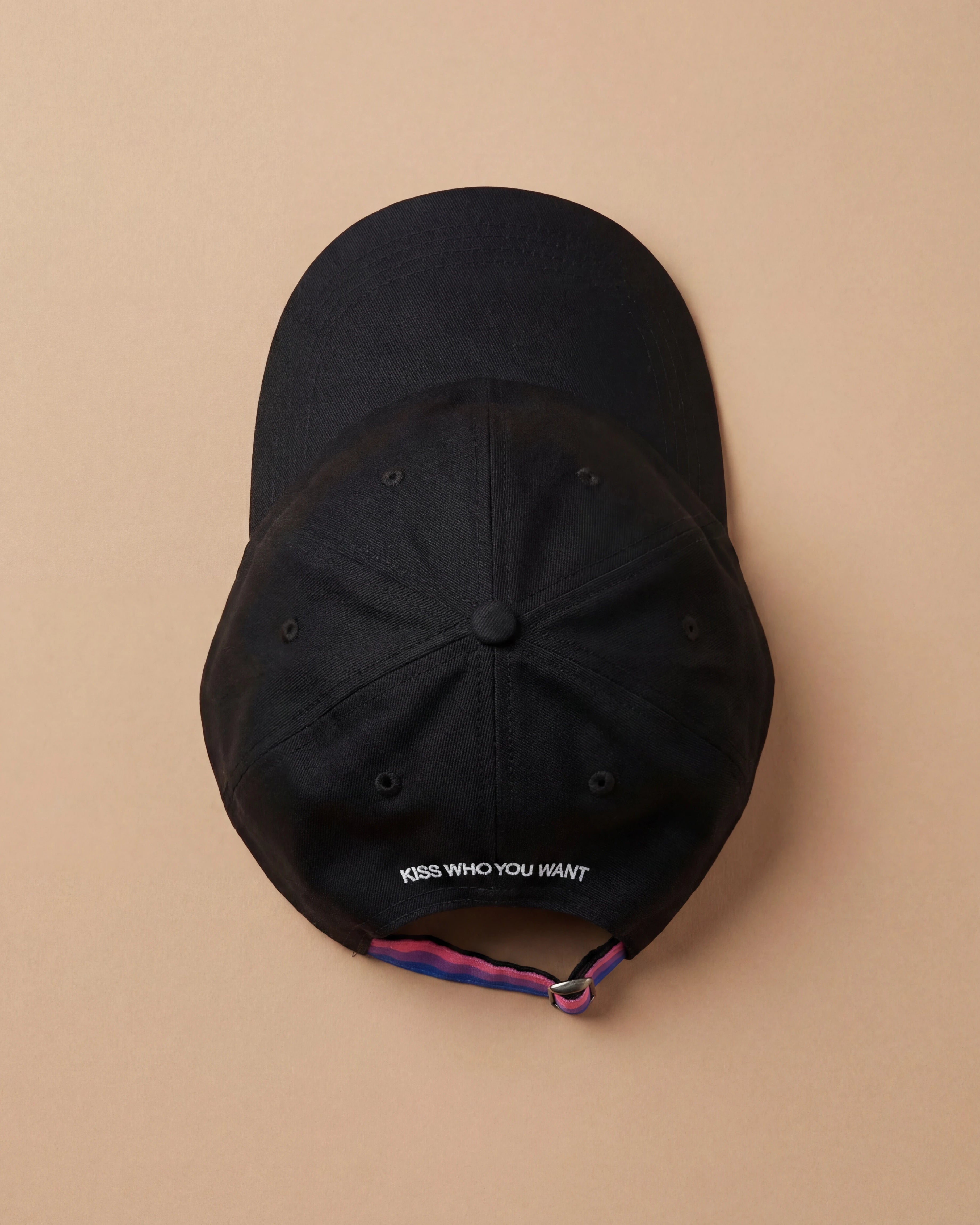 Black Biweekly cap with a strap in the colors of the bi pride flag and the text ‘Kiss Who You Want’ on the back, laying flat on beige background