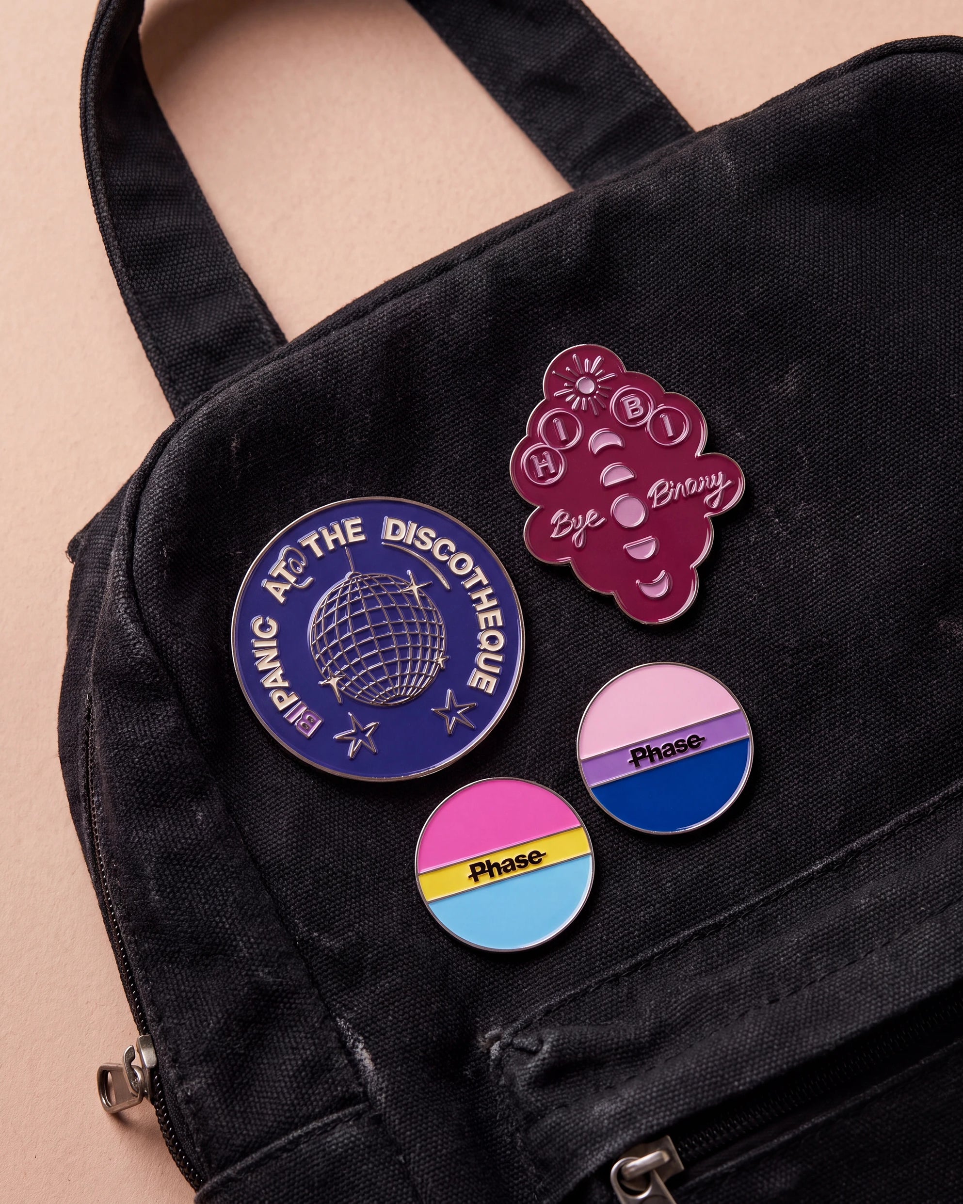 Burgundy 'Hi Bi, Bye Binary' pin, bi coloured 'Phase' pin, pan coloured 'Phase' pin and blue 'Bipanic at the discotheque' pin are displayed. All four pins are placed on a black bag.