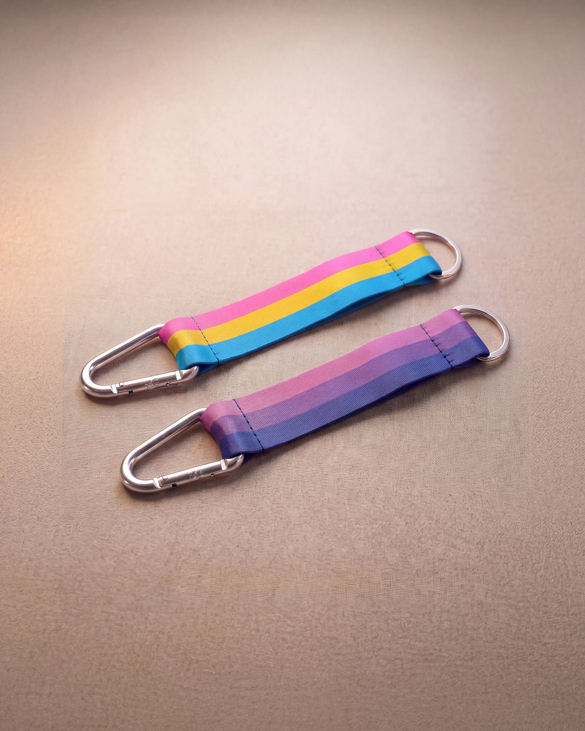 Two Keychain in bi pride and pan pride flag colors with silver carabiner and blue yarn laying next to eachother against beige background