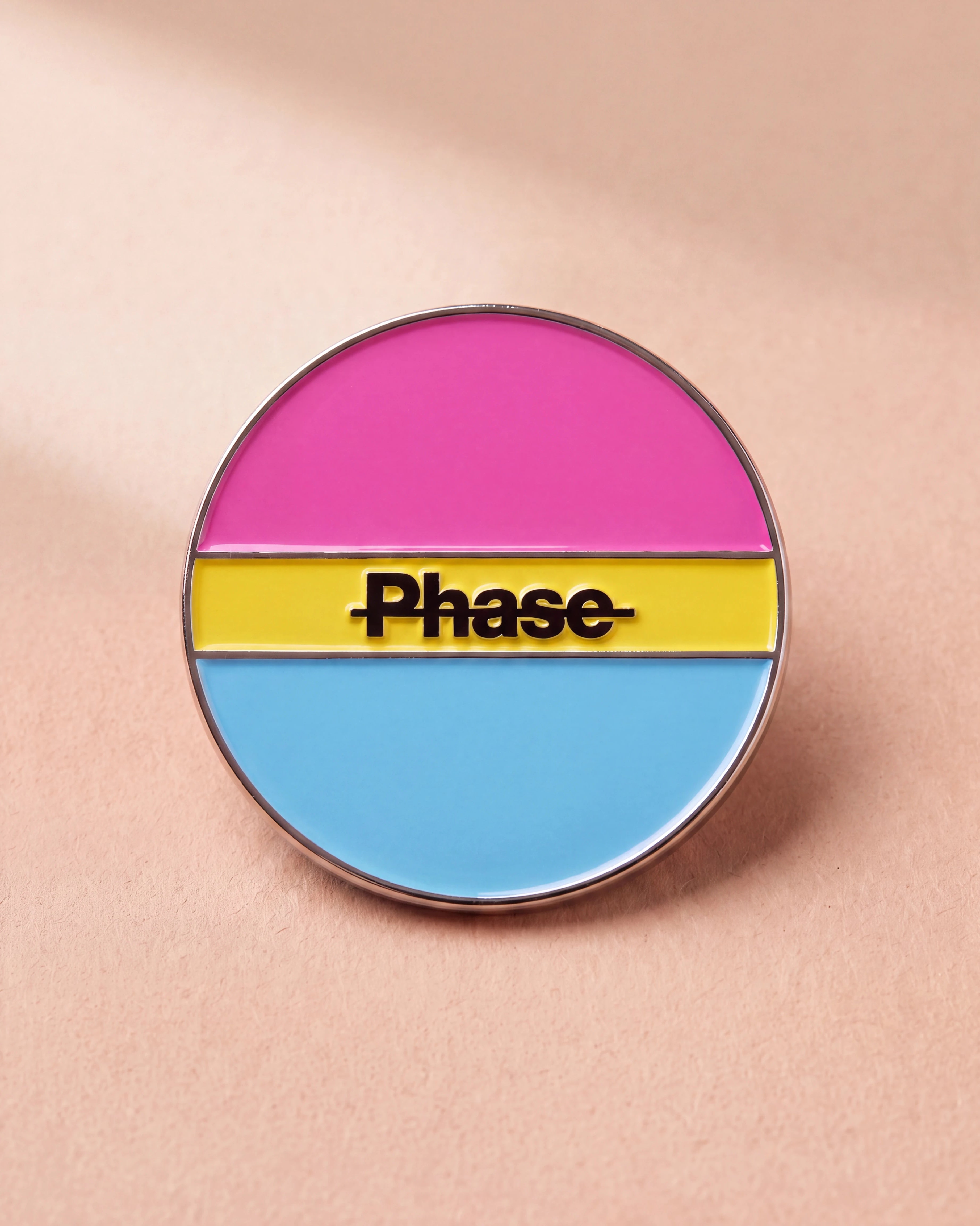 Biweekly Pin in pan colours with a crossed out 'Phase' text, flatlay against beige background