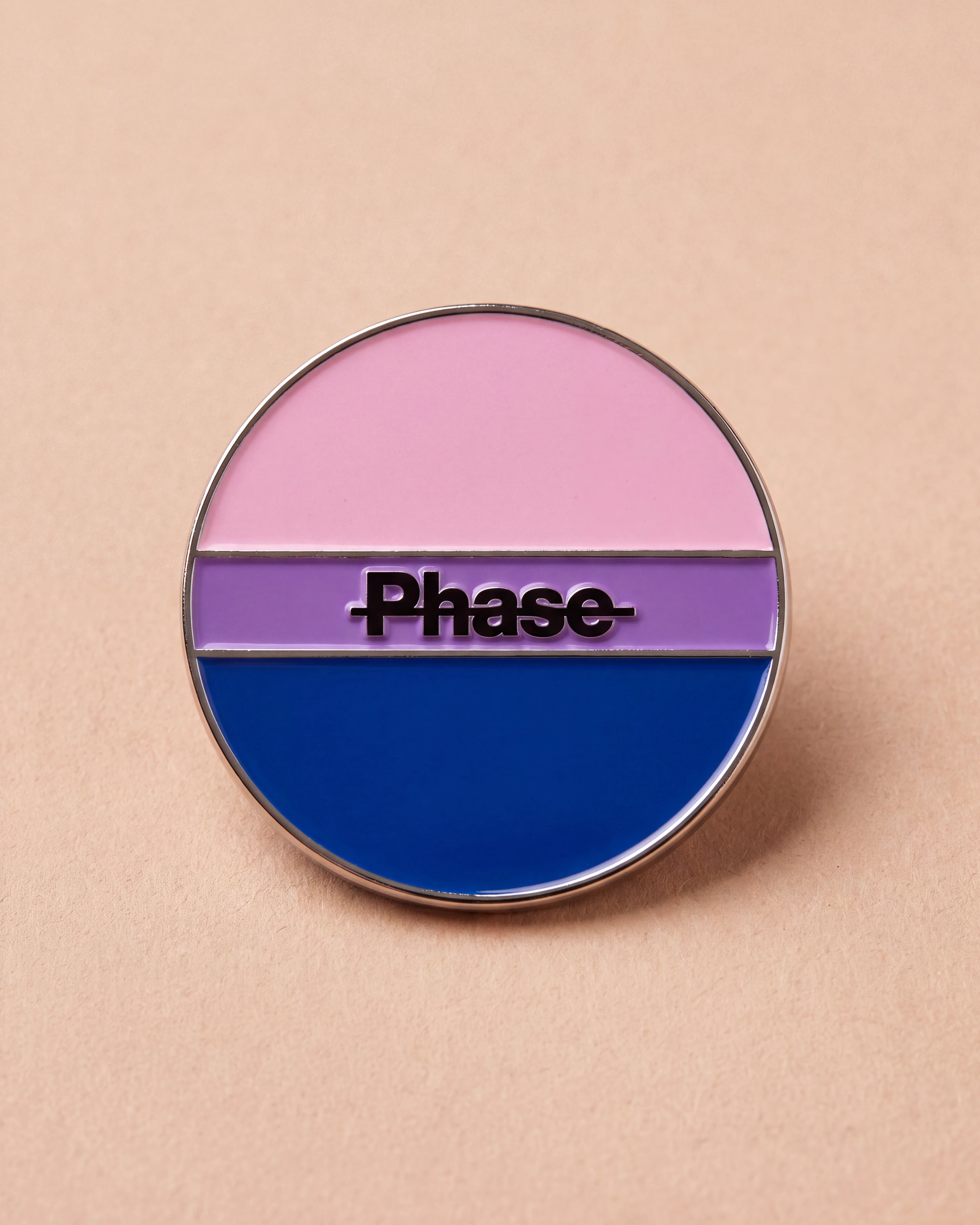 Biweekly Pin in bi colours with a crossed out 'Phase' text, flatlay against beige background