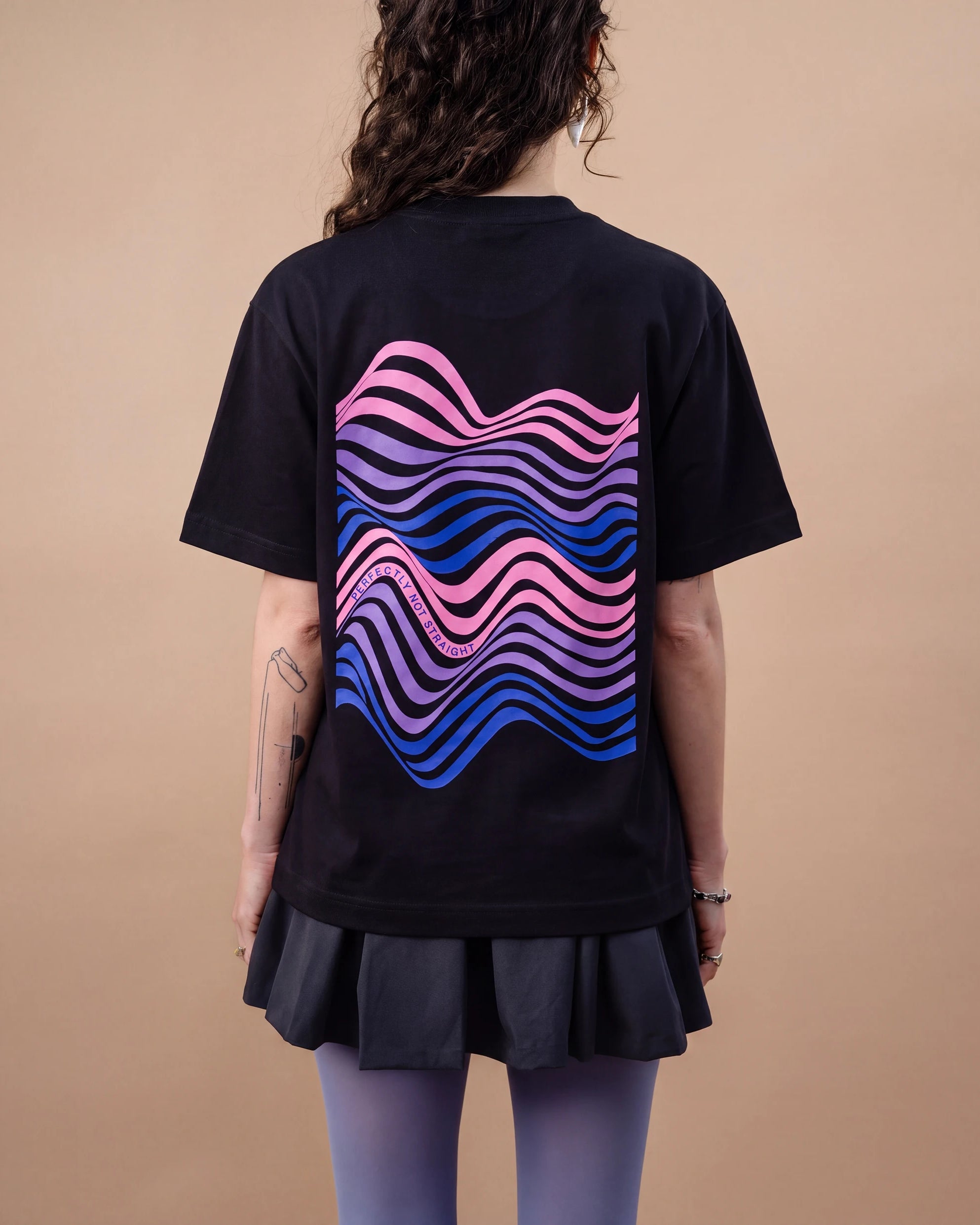 Model wears an oversized black Biweekly T-shirt with wavy bi pride flag graphic print and `Perfectly Not Straight` text on the print, back view against a beige background