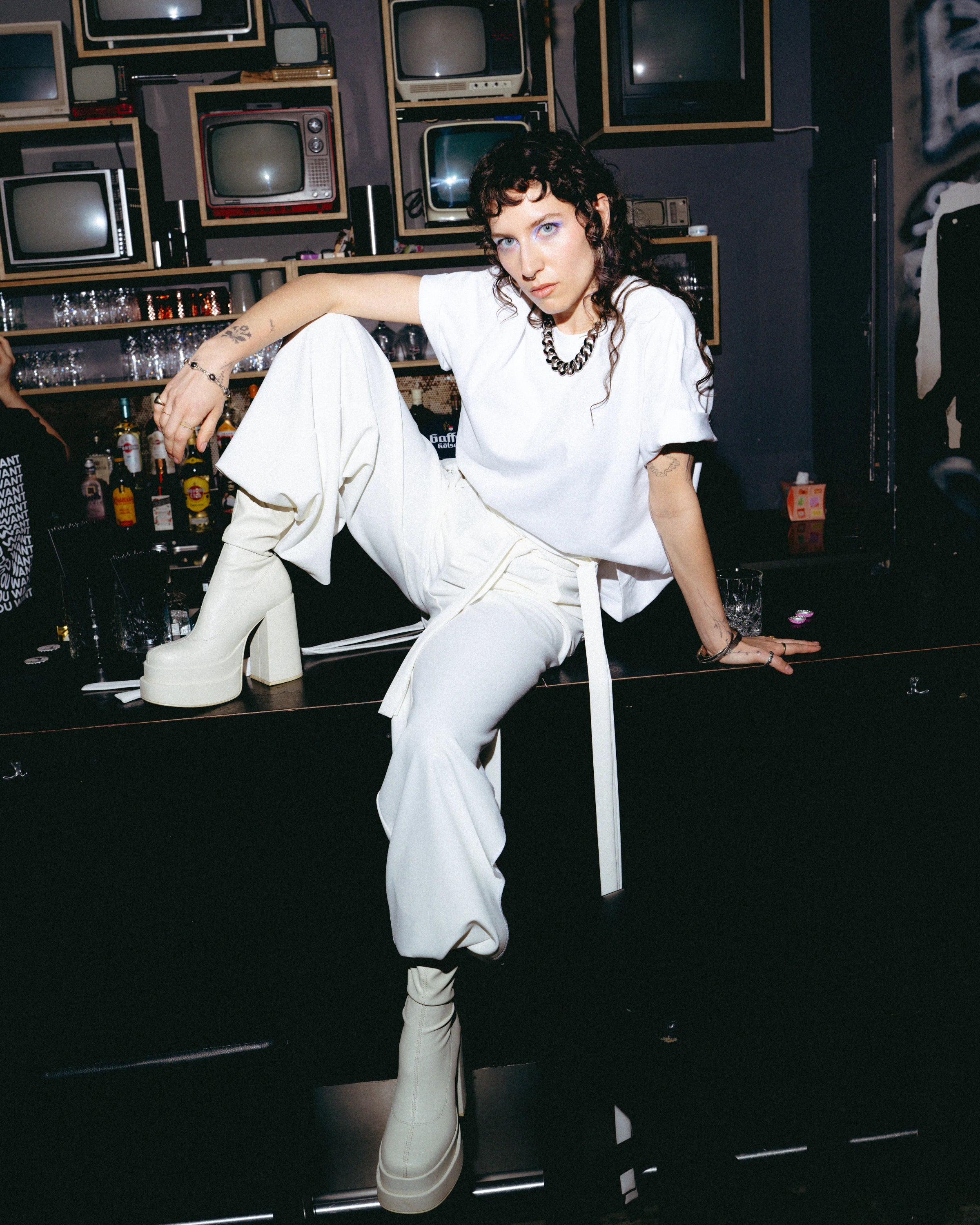 Model wearing an oversized white Biweekly T-shirt, sitting on a bar counter in a club setting