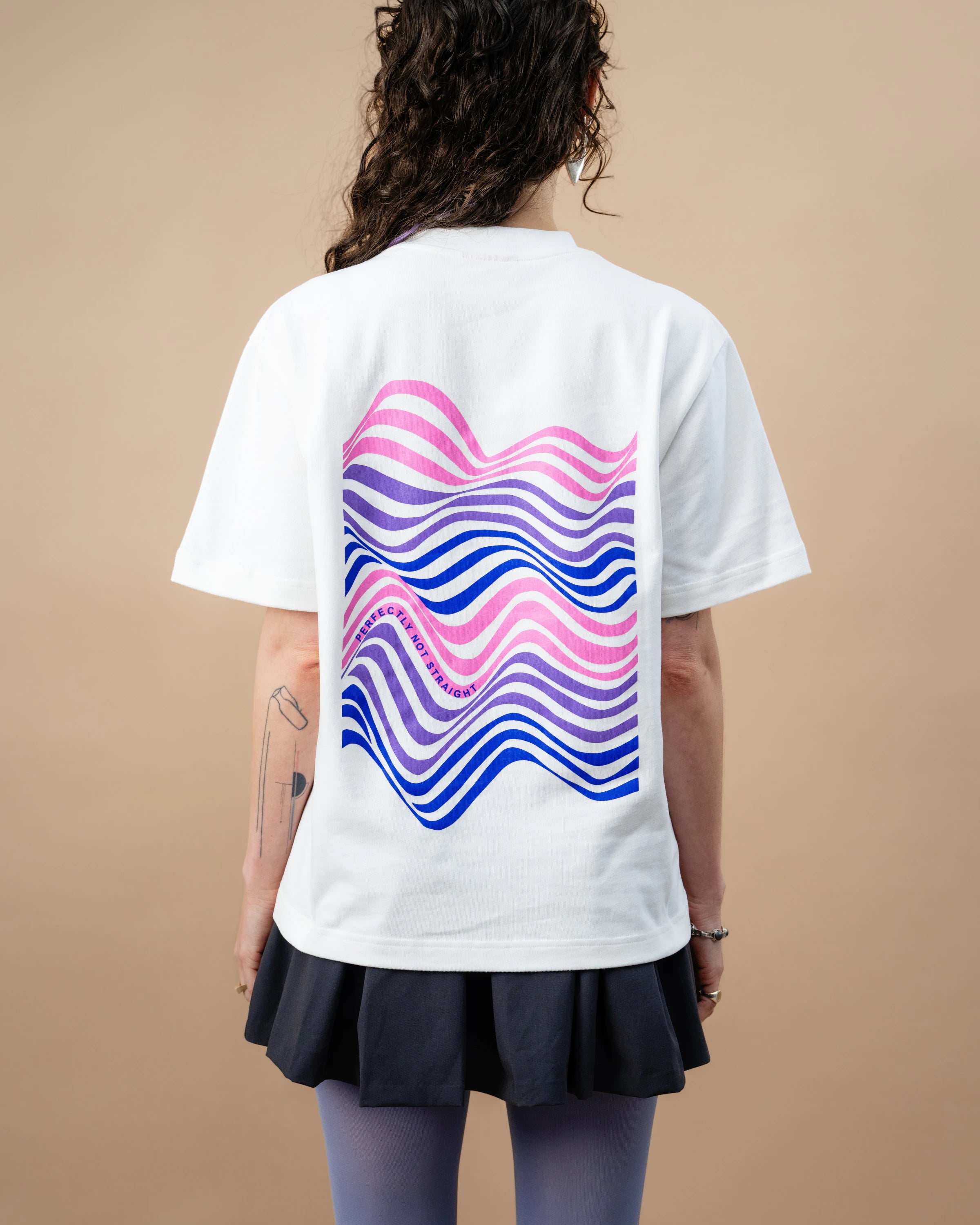 Model wears an oversized white Biweekly T-shirt with wavy bi pride flag graphic print and `Perfectly Not Straight` text on the print, back view against a beige background