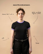 Model wearing a black Biweekly Babytee with bi pride flag trim on the collar and sleeves and black logo in the front, front view against a beige background. Text that gives information about the top. 