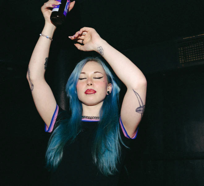Model wearing a black Biweekly Babytee with bi pride flag trim on the collar and sleeves and black logo in the front, front view in a club scene, dancing, with beer in her hand