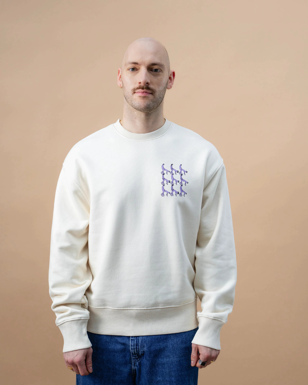 Model wearing a light beige Biweekly crewneck with graphic print of stylized figures in playful poses on the chest, front view against a beige background