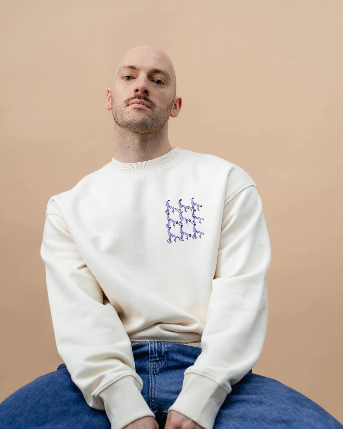 Model sitting on a chair wearing a light beige Biweekly crewneck with graphic print of stylized figures in playful poses on the chest, front view against a beige background