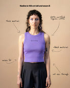 Model wearing a lilac Biweekly tank top with black ‘Assume nothing’ text, front view against a beige background. Text that gives information about the top. 