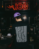 Model wearing a black oversized Biweekly T-shirt with distorted repeated ‘Kiss Who You Want’ text print, back view in a club scenario
