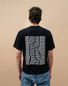 Model wearing a black oversized Biweekly T-shirt with distorted repeated ‘Kiss Who You Want’ text print, back view against a beige background