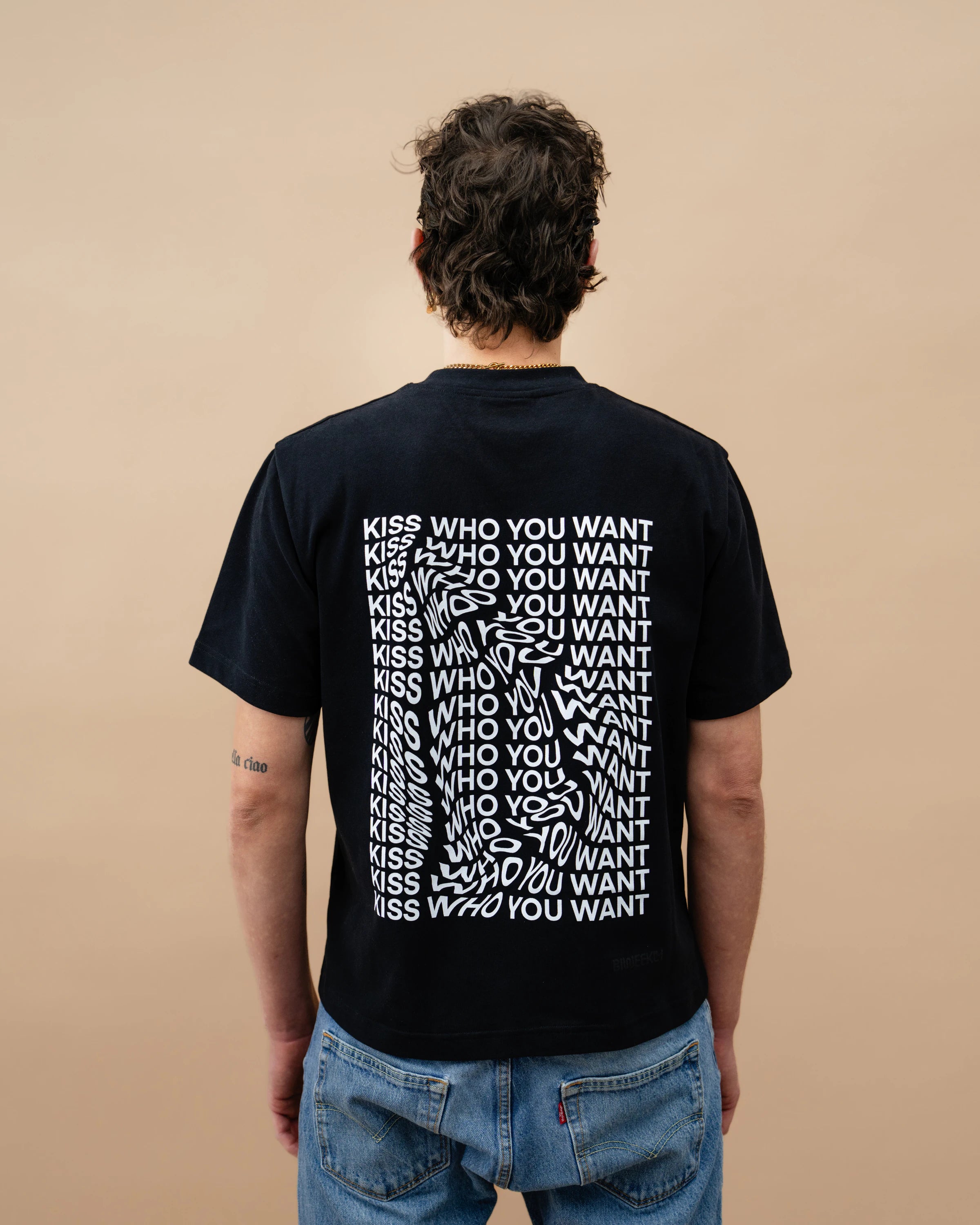 Model wearing a black oversized Biweekly T-shirt with distorted repeated ‘Kiss Who You Want’ text print, back view against a beige background