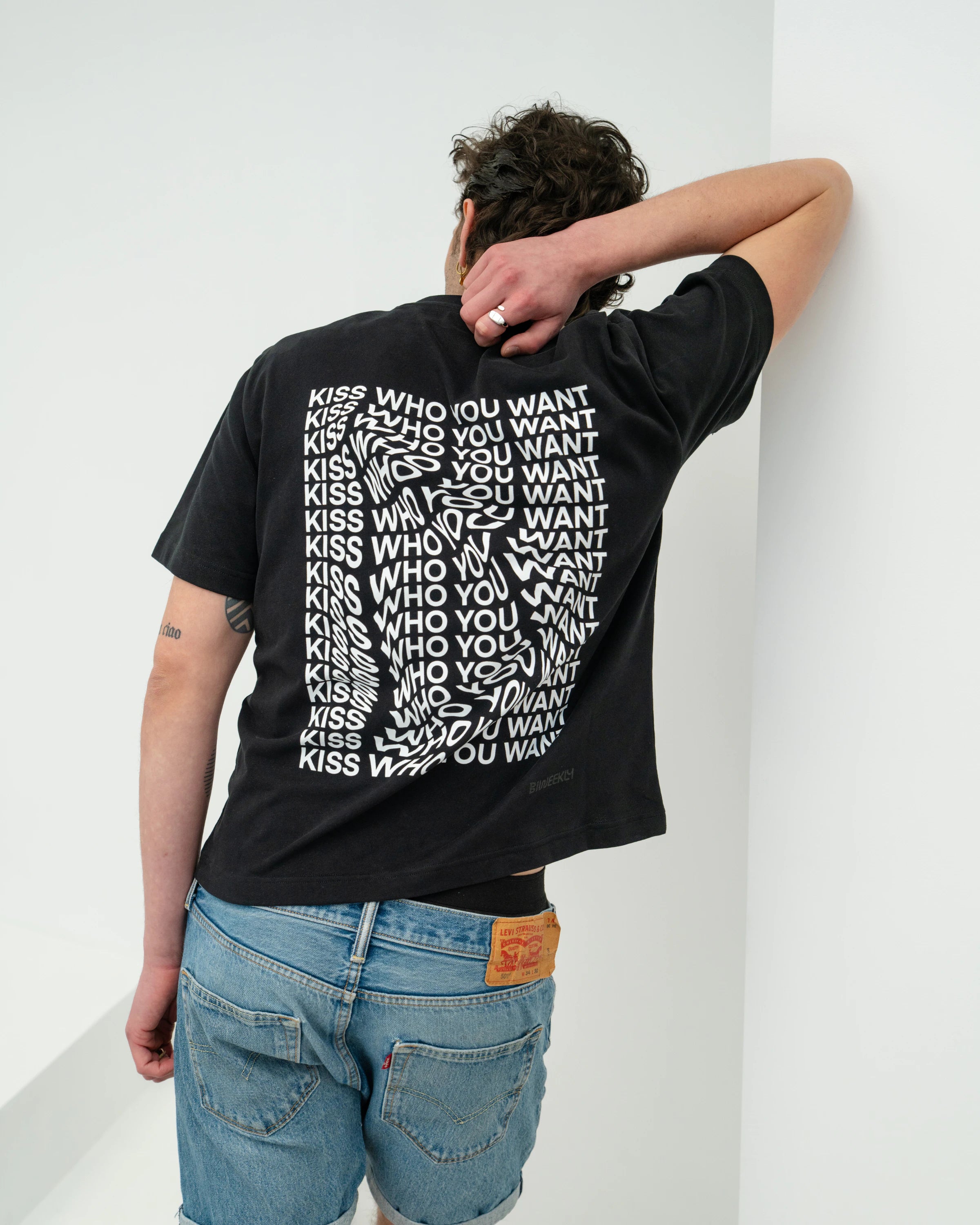 Model wearing a black oversized Biweekly T-shirt with distorted repeated ‘Kiss Who You Want’ text print, back view against a white background
