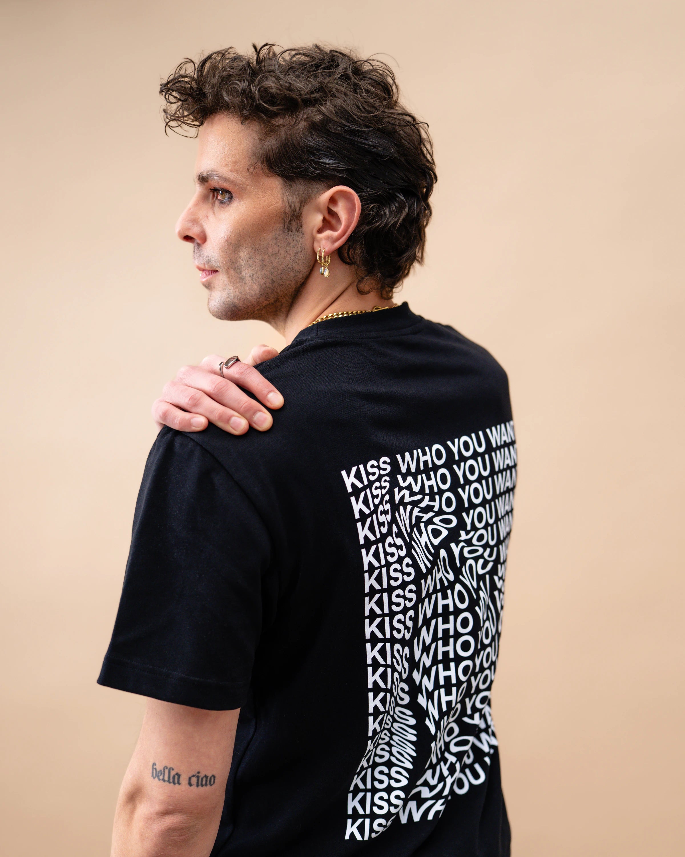 Model wearing a black oversized Biweekly T-shirt with distorted repeated ‘Kiss Who You Want’ text print, side view against a beige background