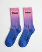 Biweekly socks with watercolor fade from pink to blue with a crossed out "Phase" on the inside and Biweekly logo on the bottom laying flat on a white background