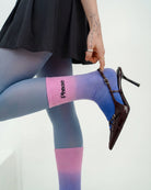 Model wearing Biweekly socks with watercolor fade from pink to blue with a crossed out "Phase" on the inside, paired with black heels, holding the foot up with a finger on the shoe, against a white background