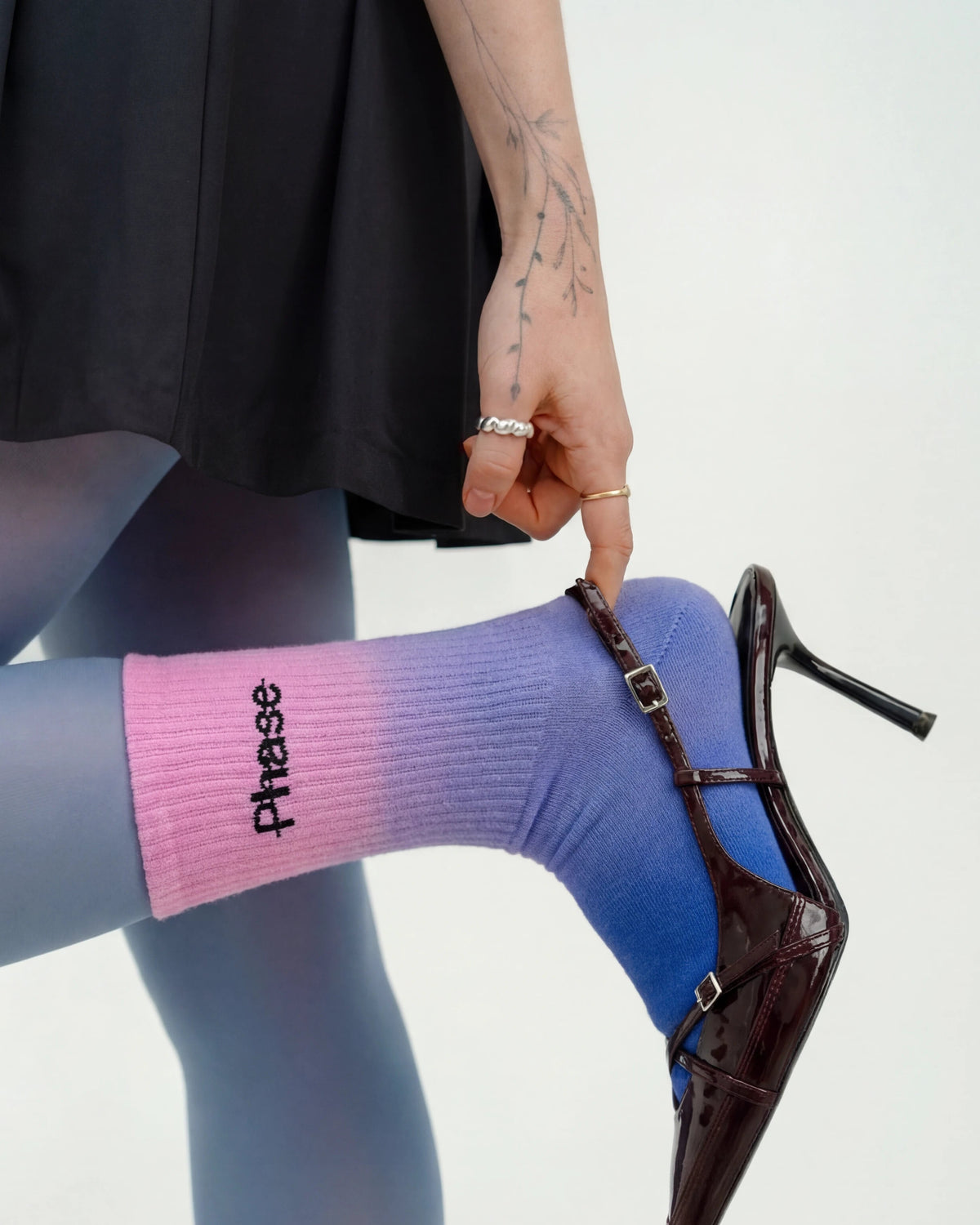 Model wearing Biweekly socks with watercolor fade from pink to blue with a crossed out "Phase" on the inside, paired with black heels, holding the foot up with a finger on the shoe, against a white background