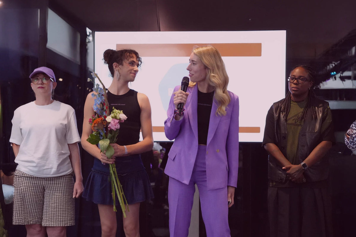 four women on a stage, one holding a microphone and one holding flowers looking at eachother