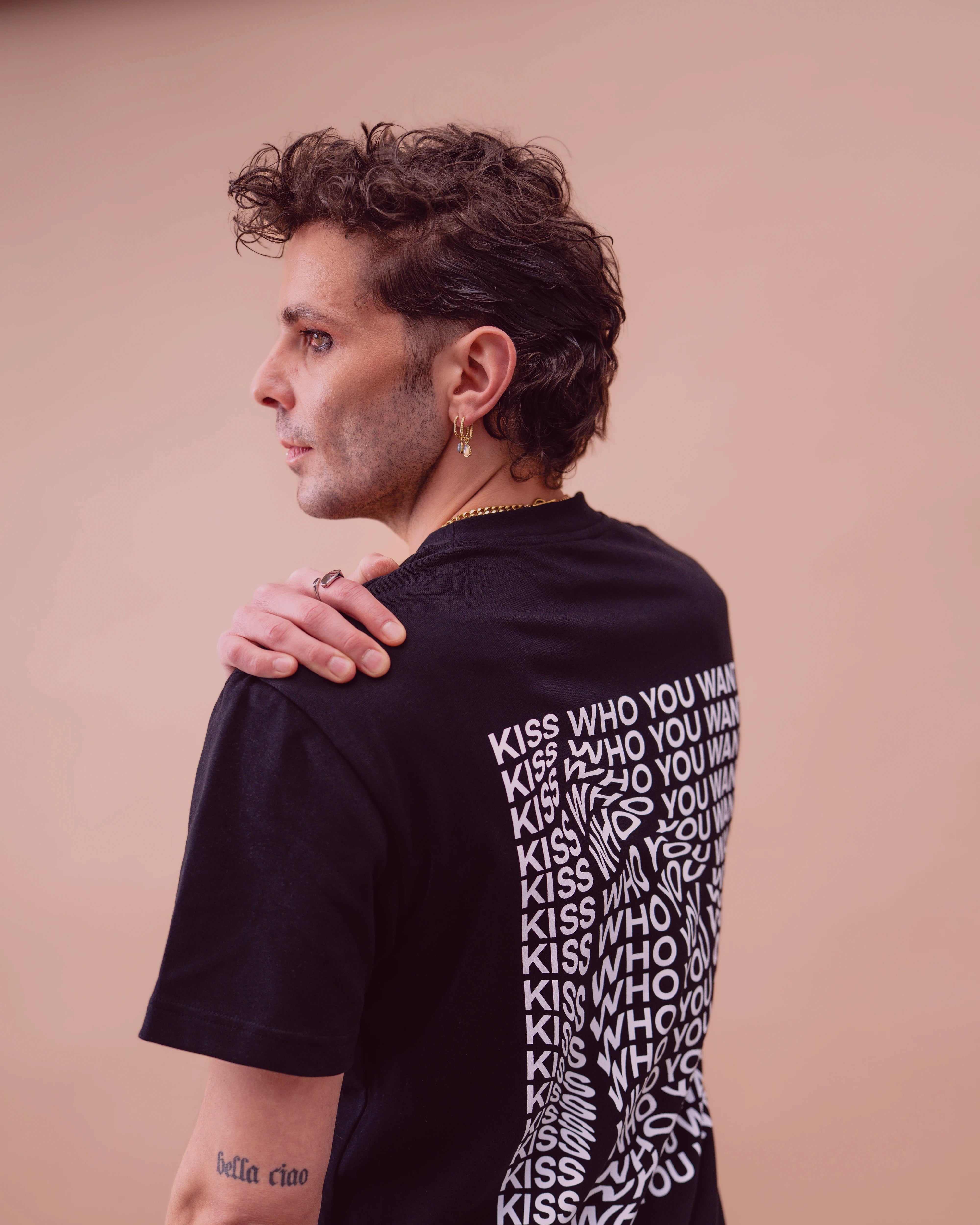 Model wearing a black oversized Biweekly T-shirt with distorted repeated ‘Kiss Who You Want’ text print, side view against a beige background with subtle burgundy overlay