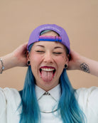 Model wearing a lilac Biweekly cap with a strap in the colors of the bi pride flag and the text ‘Kiss Who You Want’ on the back, sticking out their tongue against a beige background