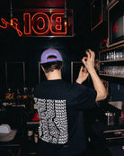 Model wearing a lilac Biweekly cap with bi pride flag strap and ‘Kiss Who You Want’ text on the back, back view, in a club scenario with a cocktail shaker in his hand
