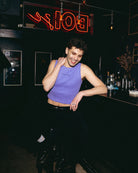 Model wearing a lilac Biweekly tank top with black ‘Assume nothing’ text, front view sitting in a chair in a cub scene