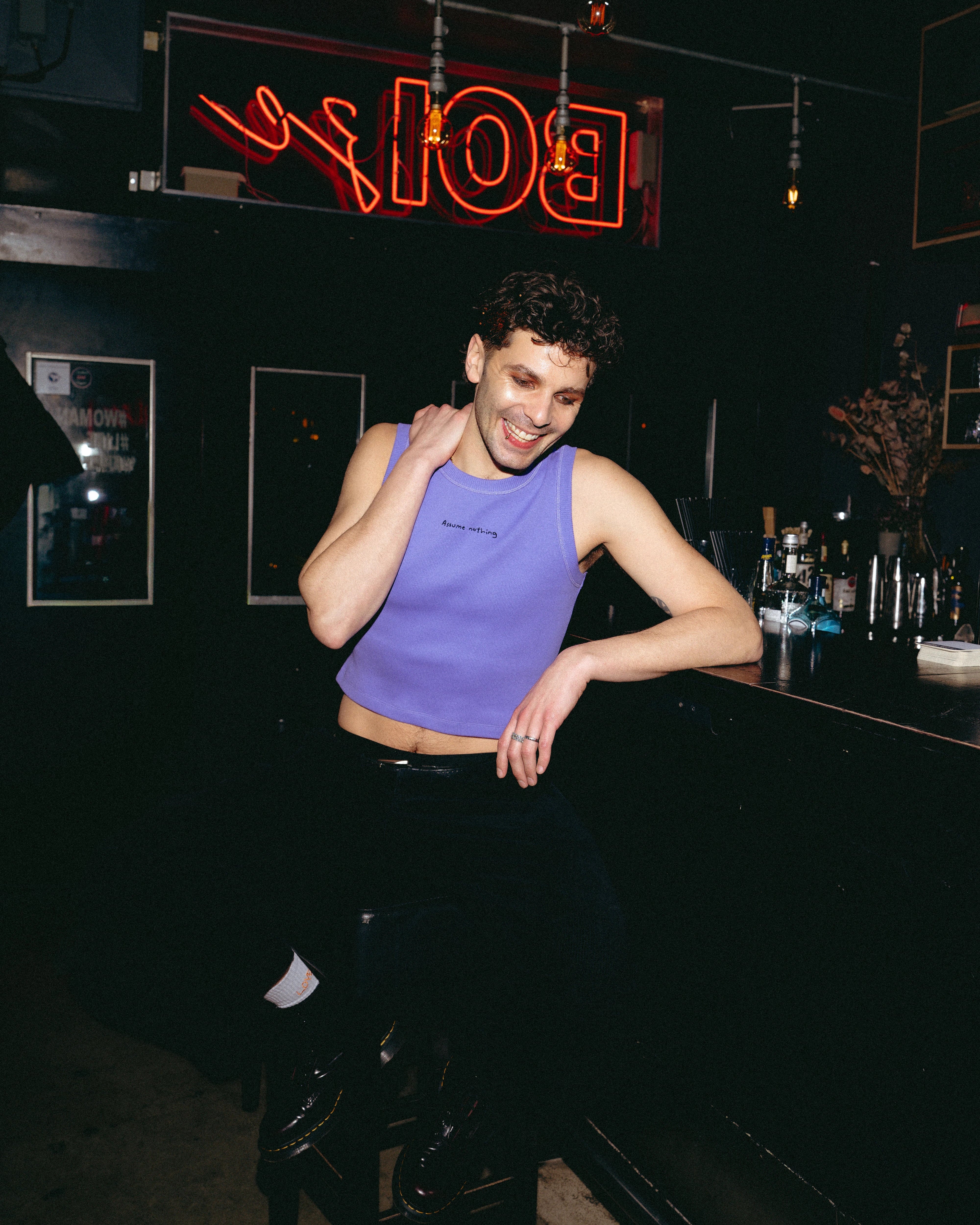 Model wearing a lilac Biweekly tank top with black ‘Assume nothing’ text, front view sitting in a chair in a cub scene