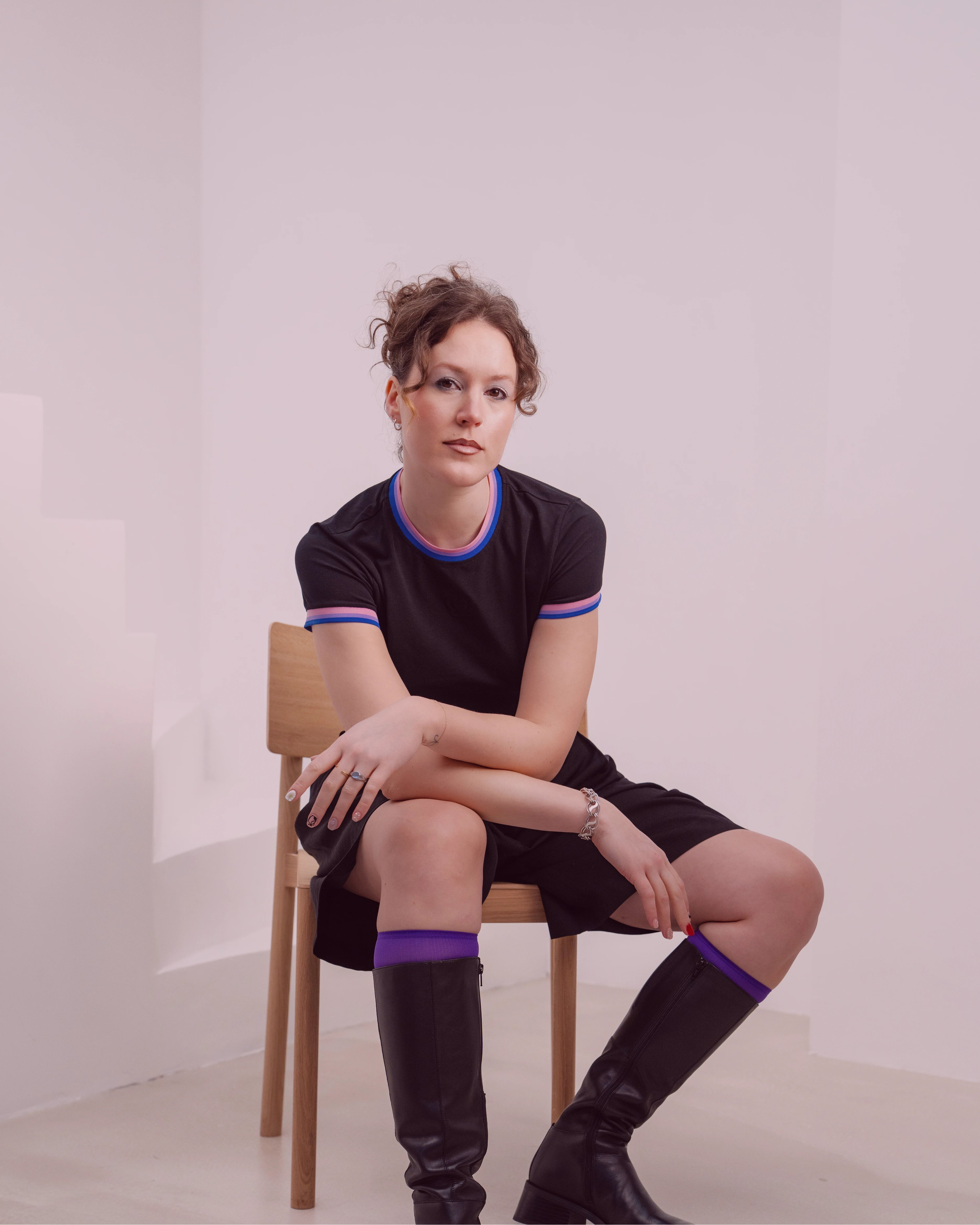 Model wearing a black Biweekly Babytee with bi pride flag trim on the collar and sleeves and black logo in the front, sitting on a chair front view against a white background with subtle burgundy color overlay