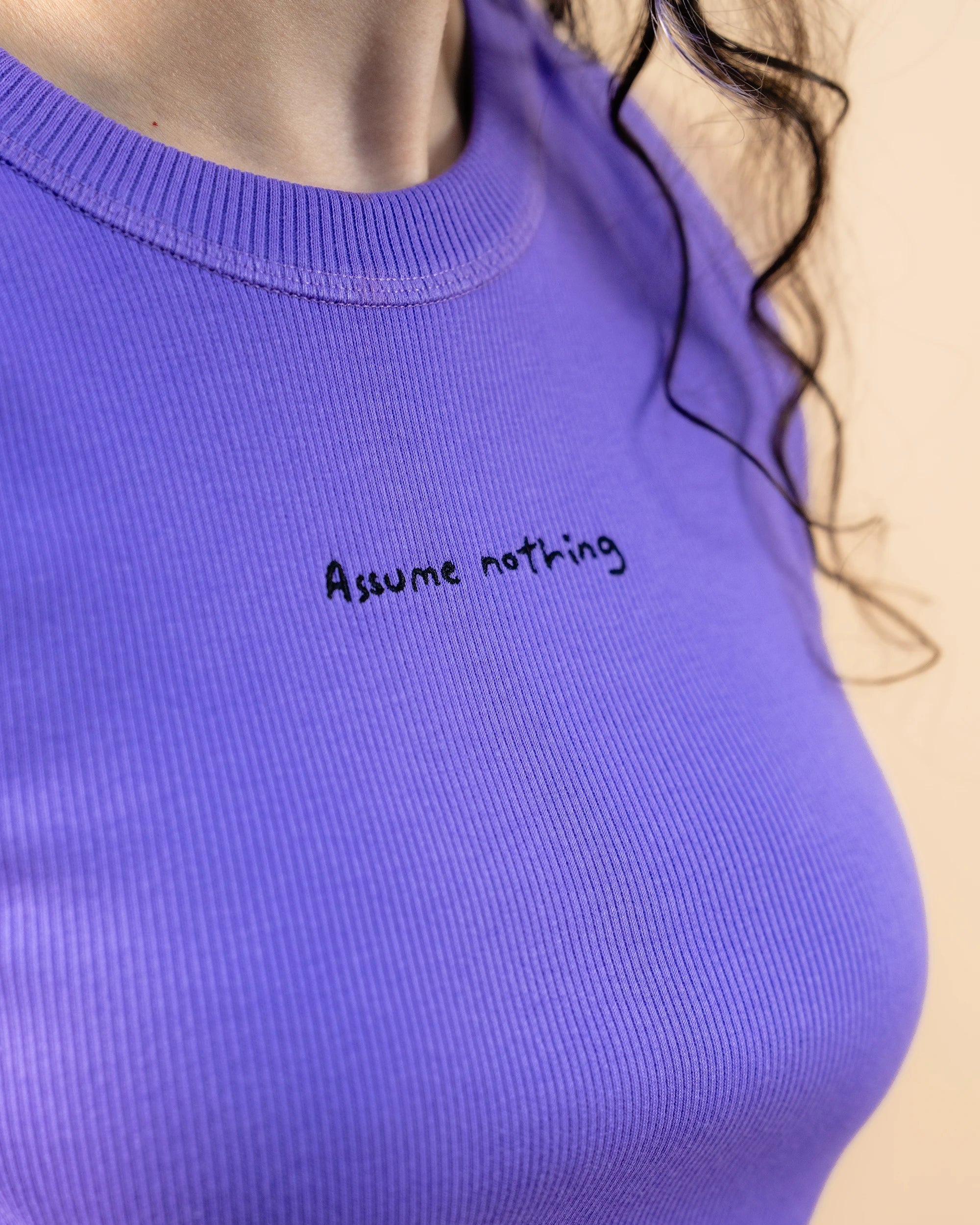 Detailshot of model wearing a lilac Biweekly tank top with black ‘Assume nothing’ text, side view against a beige background