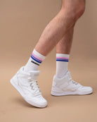 Model wearing white Biweekly socks with bi pride flag stripes and text logo underneath, paired with white sneakers, against a beige background