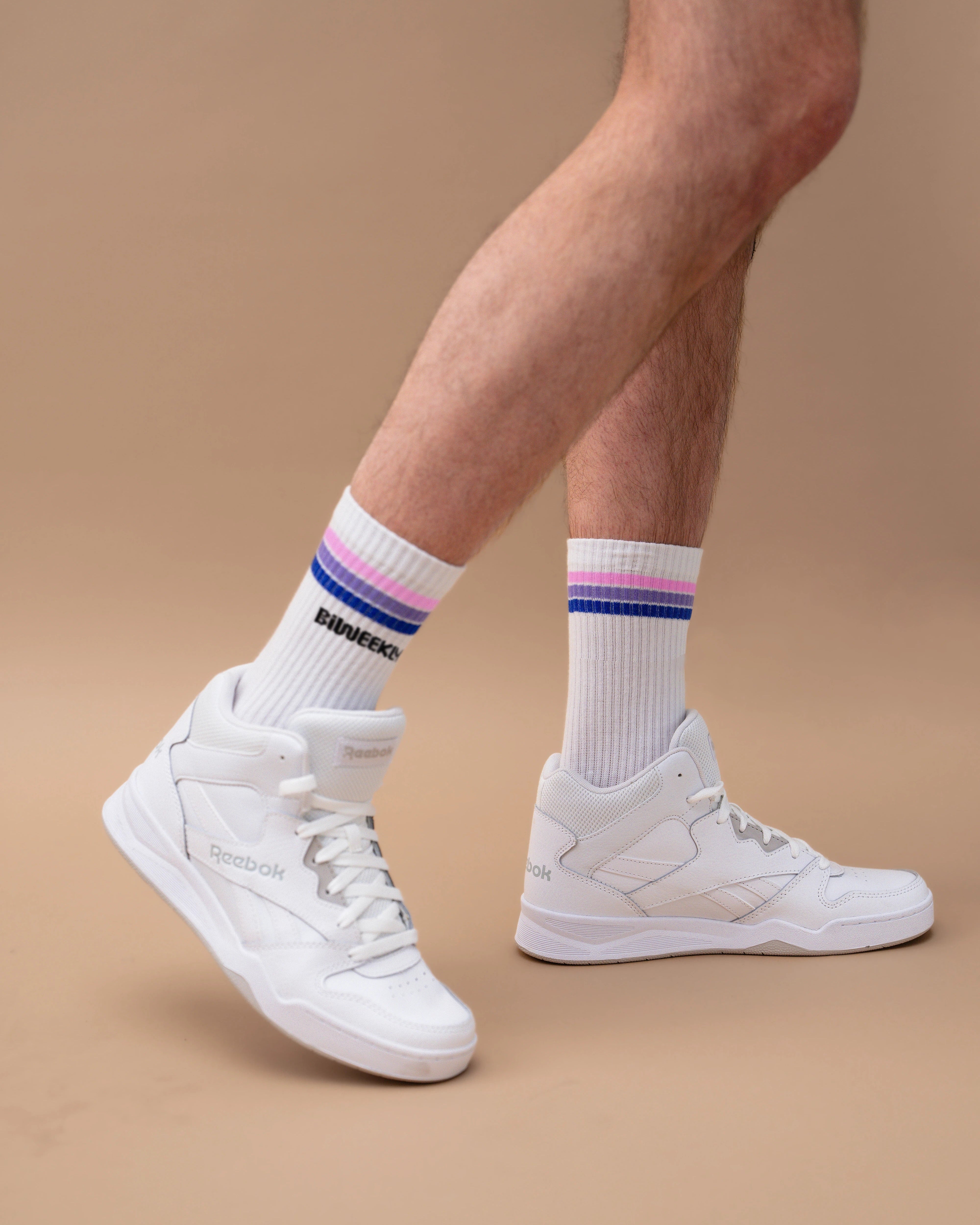 Model wearing white Biweekly socks with bi pride flag stripes and text logo underneath, paired with white sneakers, against a beige background