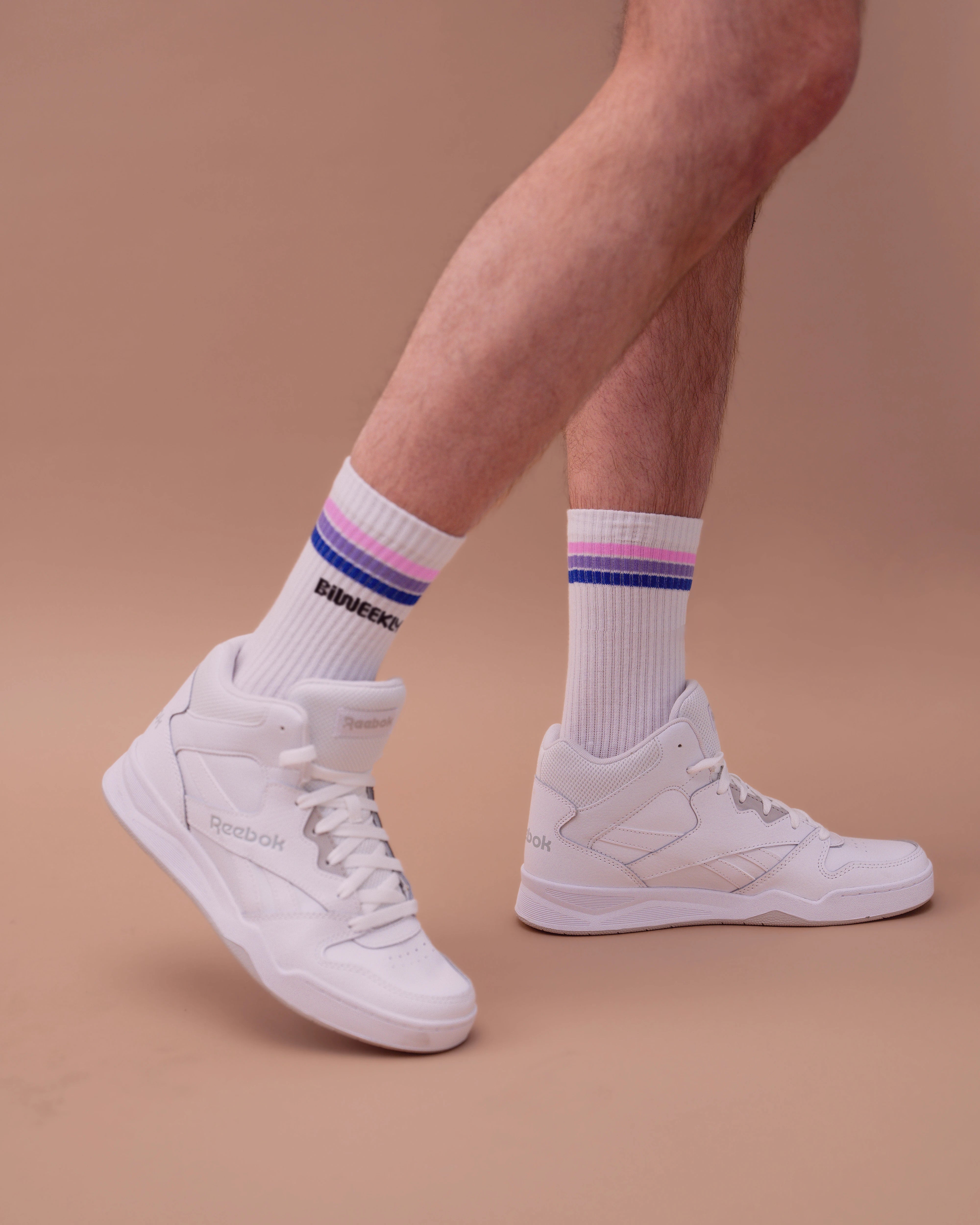 Model wearing white Biweekly socks with bi pride flag stripes and text logo underneath, paired with white sneakers, against a beige background
