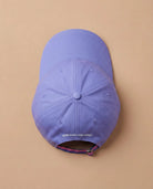 Lilac Biweekly cap with a strap in the colors of the bi pride flag and the text ‘Kiss Who You Want’ on the back, laying flat on beige background