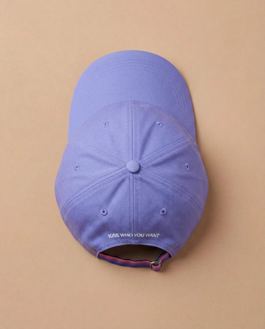 Lilac Biweekly cap with a strap in the colors of the bi pride flag and the text ‘Kiss Who You Want’ on the back, laying flat on beige background