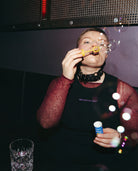 Model wearing a black Biweekly tank top with lilac ‘Kiss Who You Want’ text, blowing bubbles in a club setting
