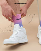 Model wearing Biweekly socks, size 42-45, with watercolor fade from pink to blue with a crossed out "Phase" on the inside, paired with white sneakers, against a beige background, with text that says it is tennis socks style and organic cotton 