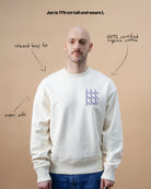 Model wearing a light beige Biweekly crewneck with graphic print of stylized figures in playful poses on the chest, front view against a beige background. Text with arrows that say relaxed boxy fit, super soft, GOTS certified organic cotton and "Jan is 179 cm tall and wears L"
