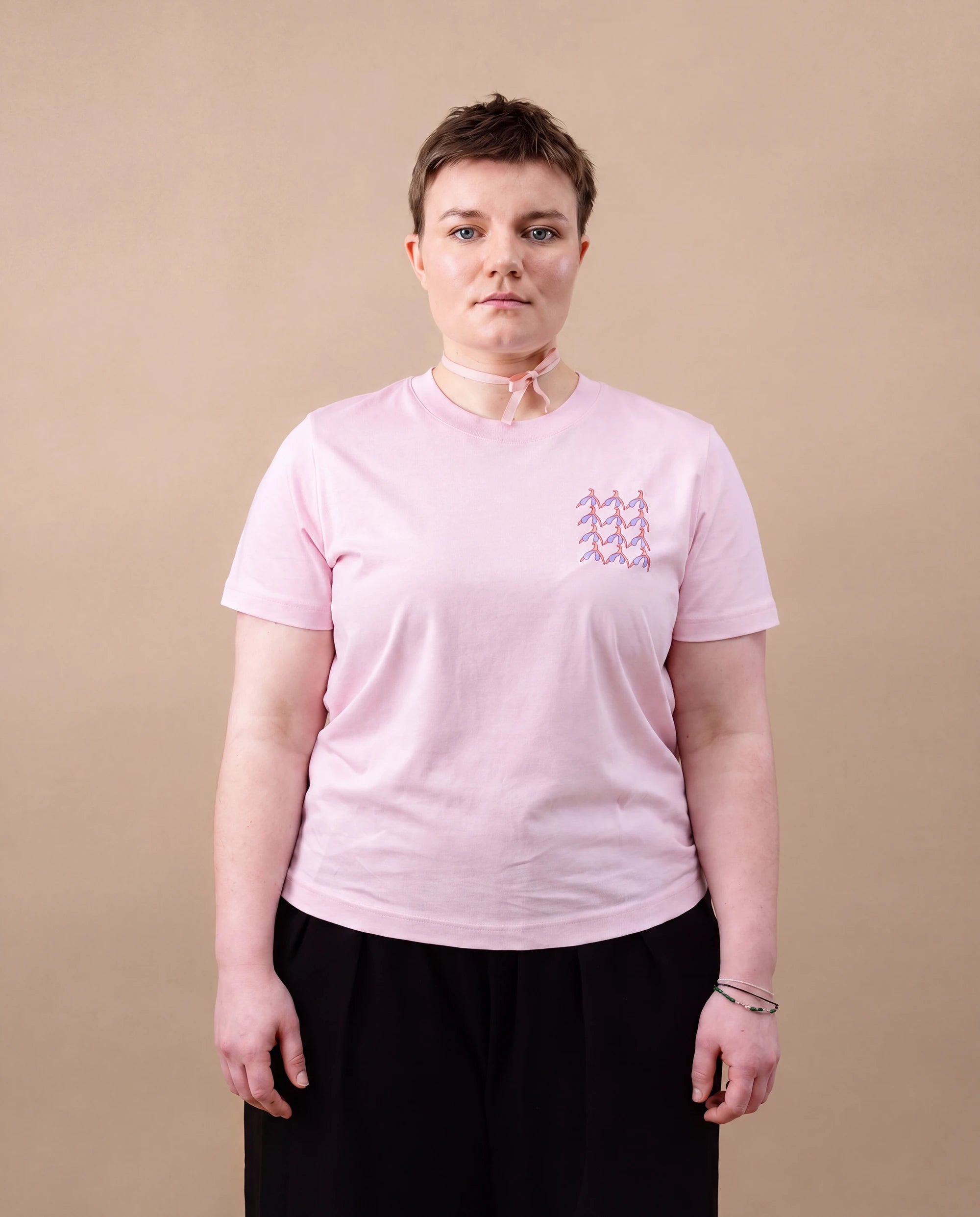 Model wearing light pink Biweekly T-shirt with clit graphic print on right chest area, front view, against beige background