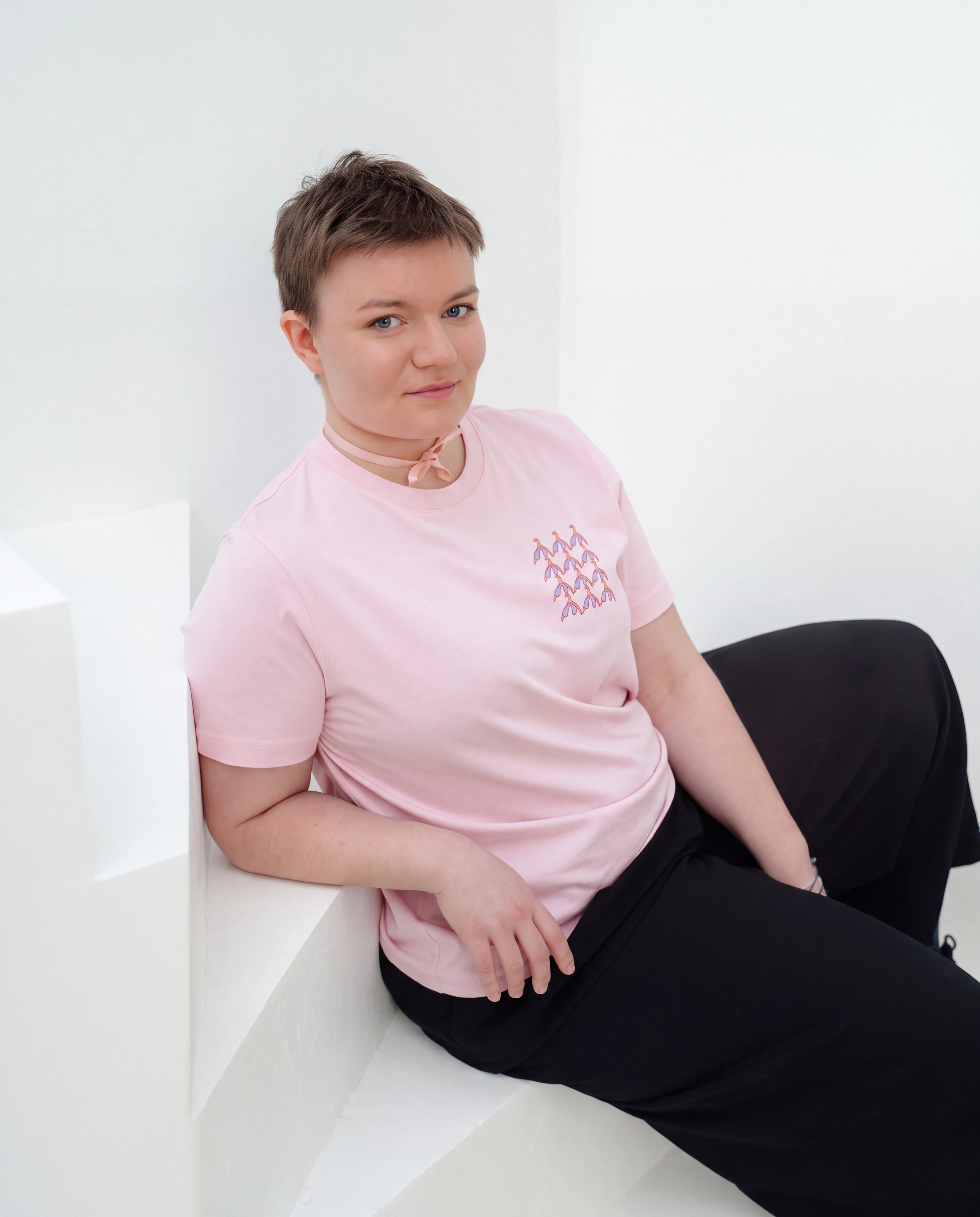 Model wearing light pink Biweekly T-shirt with clit graphic print on right chest area, front view, sitting on stairs against white background